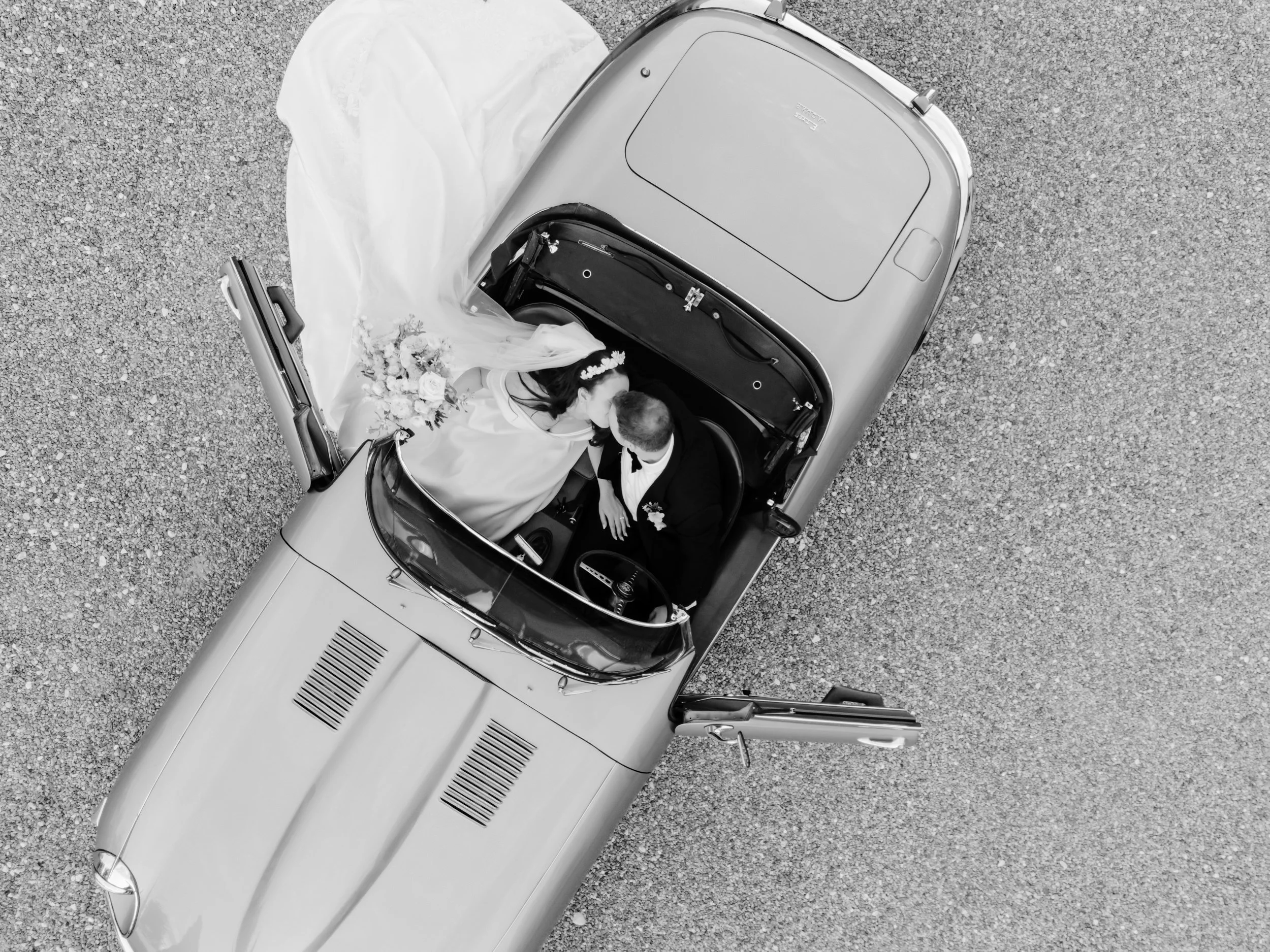 A black-and-white aerial photograph of a vintage convertible car with two people inside, a bride and groom, sitting close together. The bride is wearing a long dress and a floral crown, holding a bouquet of flowers. The groom is dressed in a suit wit