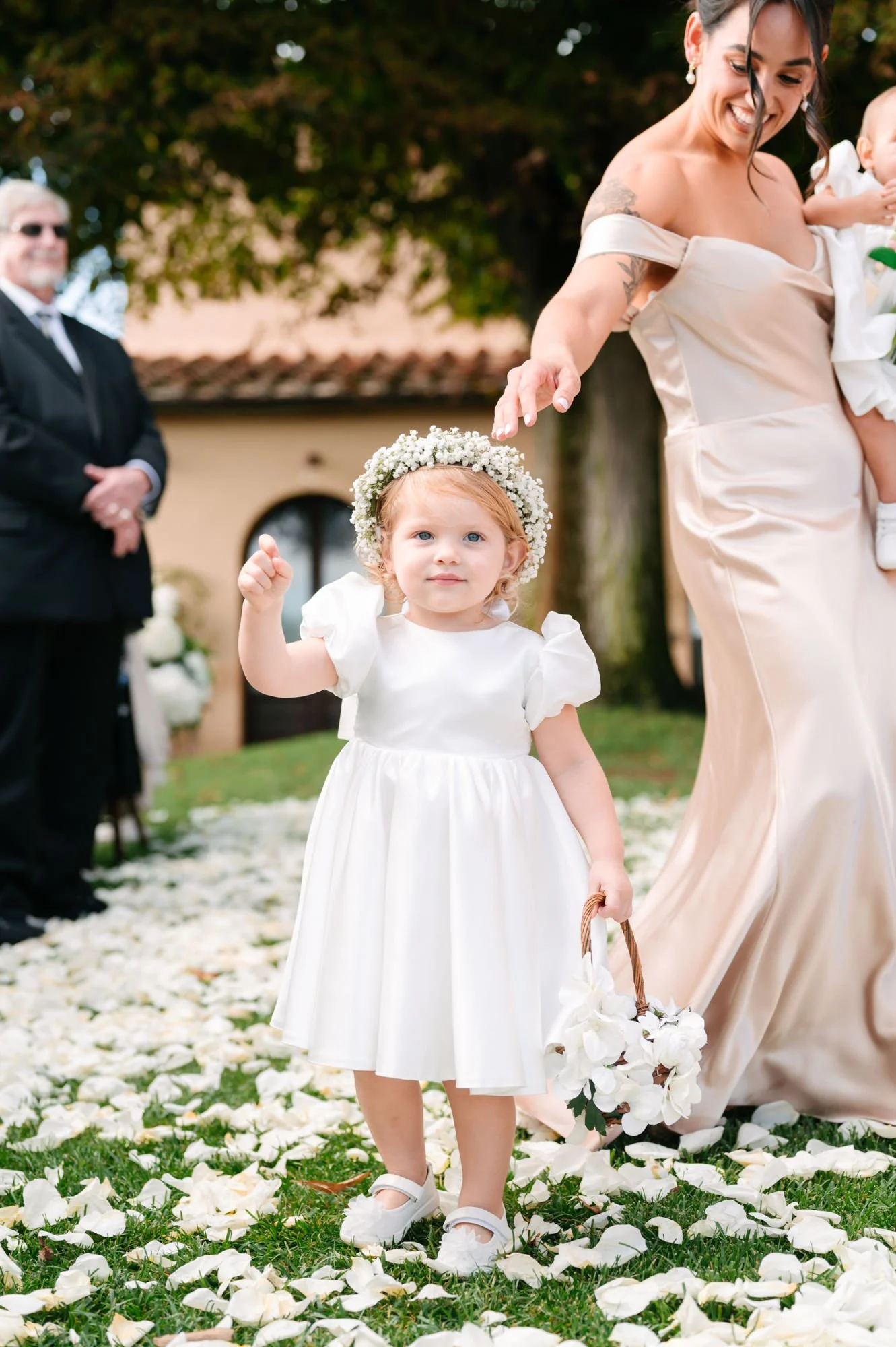 A young girl dressed in a white dress and floral crown holding a bouquet of white flowers, standing on an outdoor wedding aisle covered with white flower petals, with a woman beside her and blurred adult figures in the background.