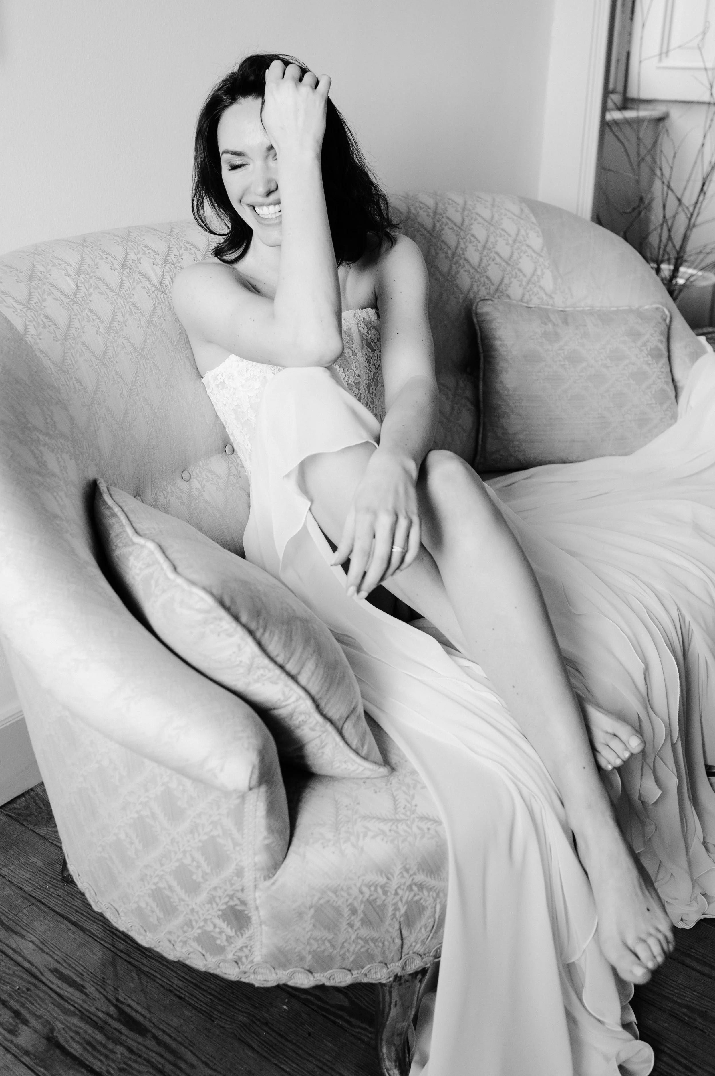 A woman with dark hair sitting on a vintage-style sofa, laughing and holding her head with one hand, wearing a light-colored dress, with her legs crossed and relaxed in a cozy indoor setting.