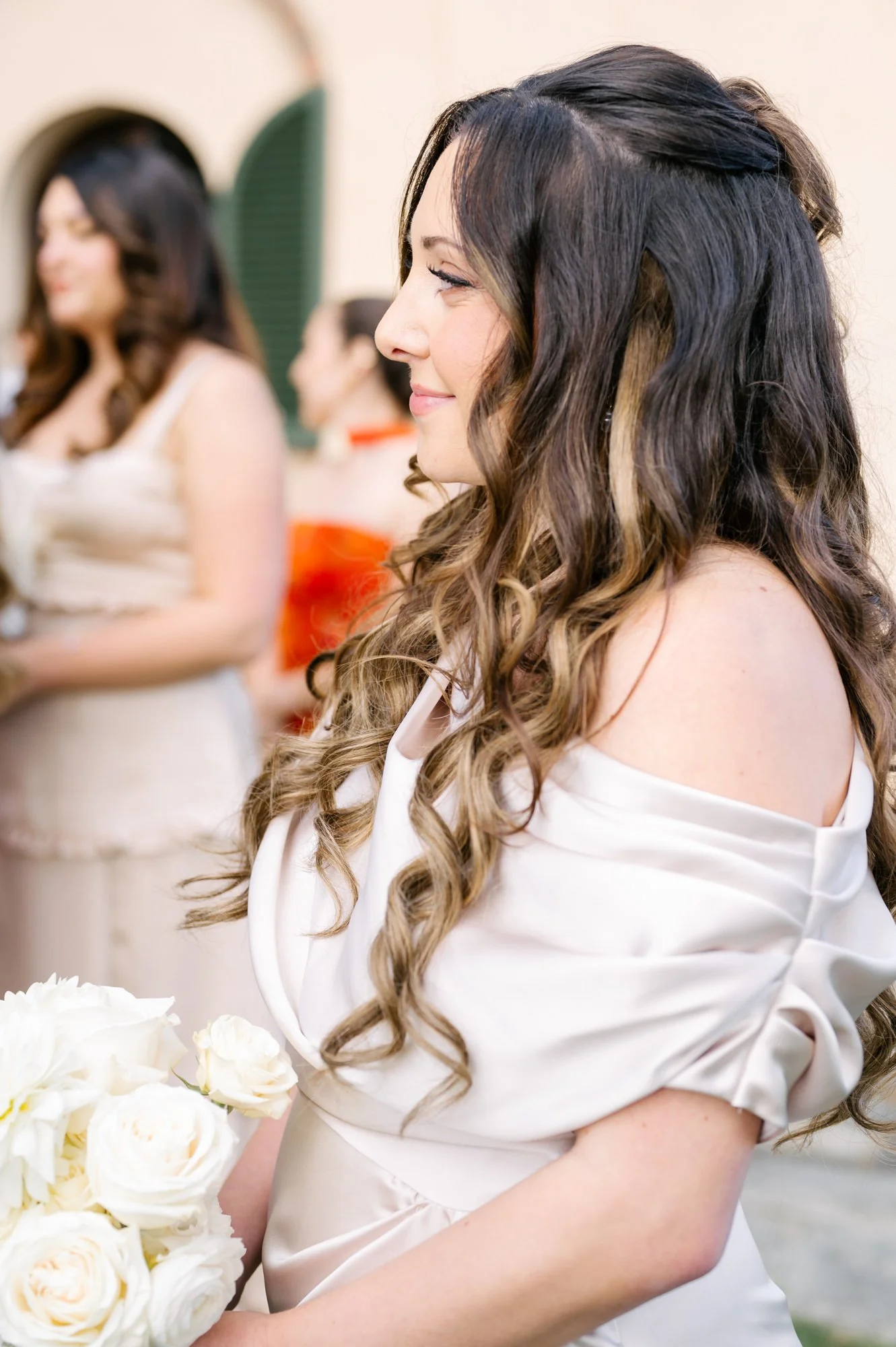 A woman with long, curly brown hair and a white dress holding a bouquet of white roses, smiling to the side. Other women in dresses are blurred in the background.