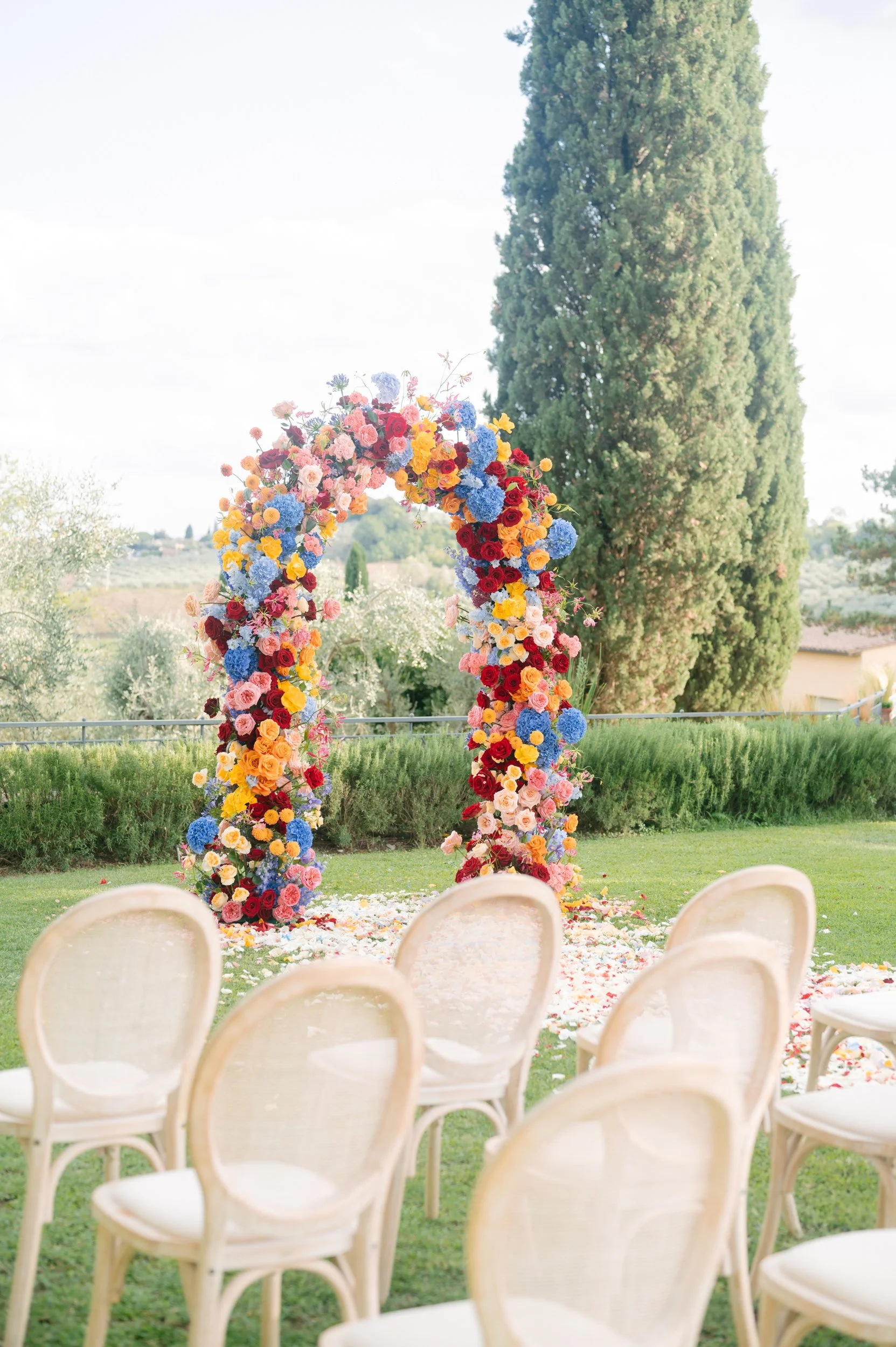 A decorated outdoor wedding ceremony setup with cream-colored chairs facing a colorful flower arch on a grassy lawn, with a large tree and a scenic landscape in the background.