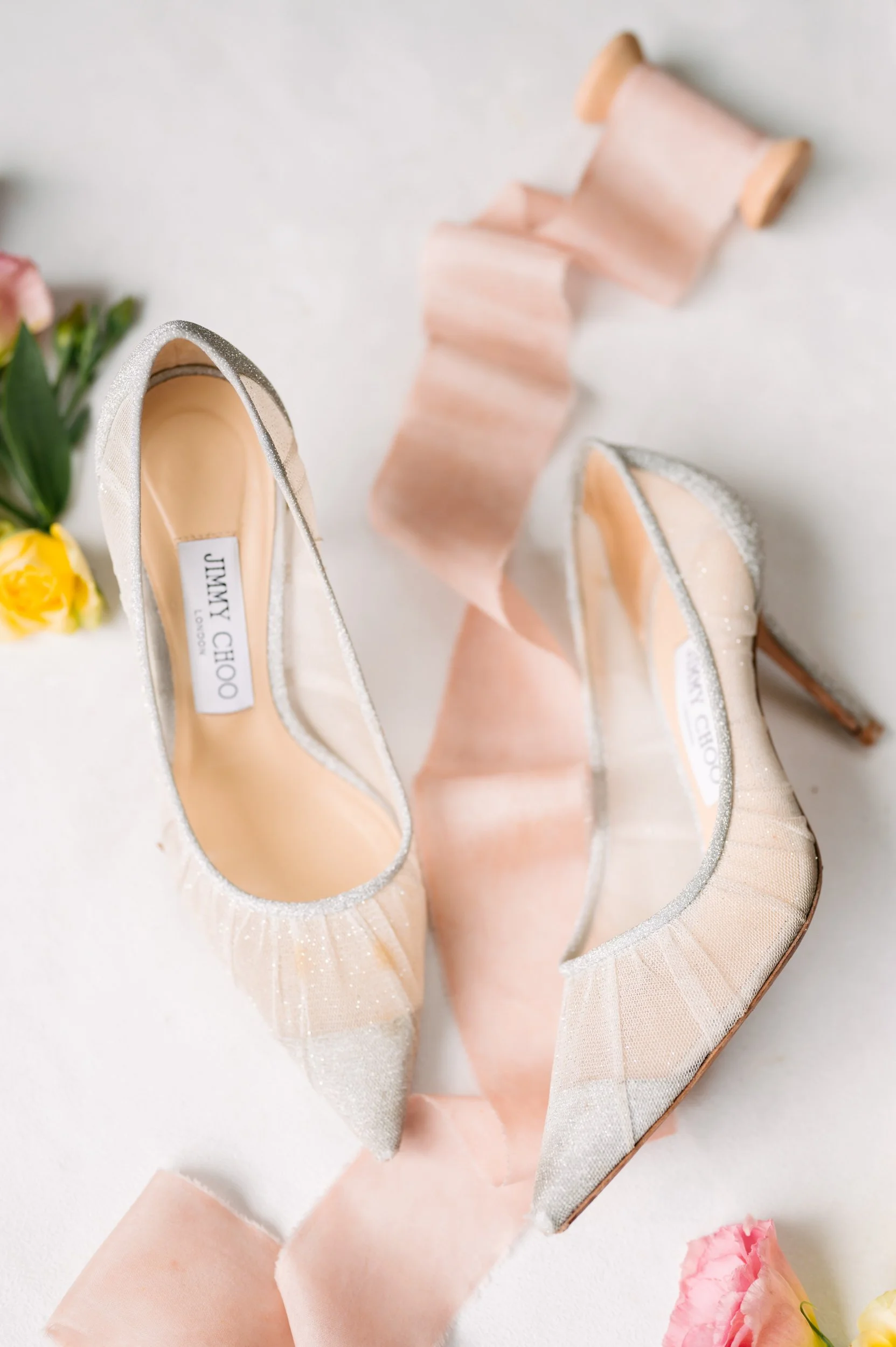 A pair of sparkling Jimmy Choo high-heeled shoes on a white surface with pink and yellow flowers and pink ribbons.