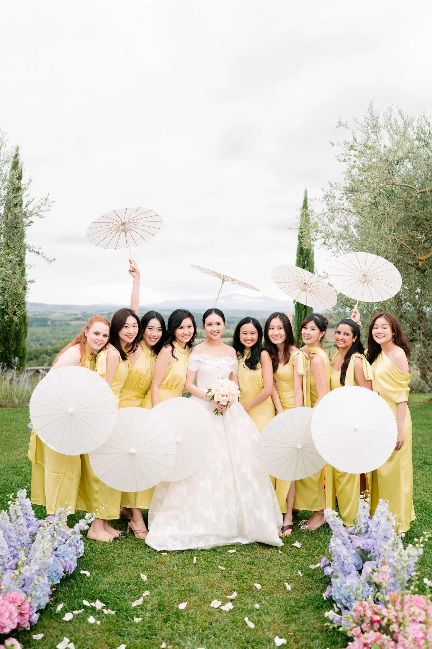 Bride in white wedding gown holding a bouquet surrounded by bridesmaids in yellow dresses holding white parasols outdoors.