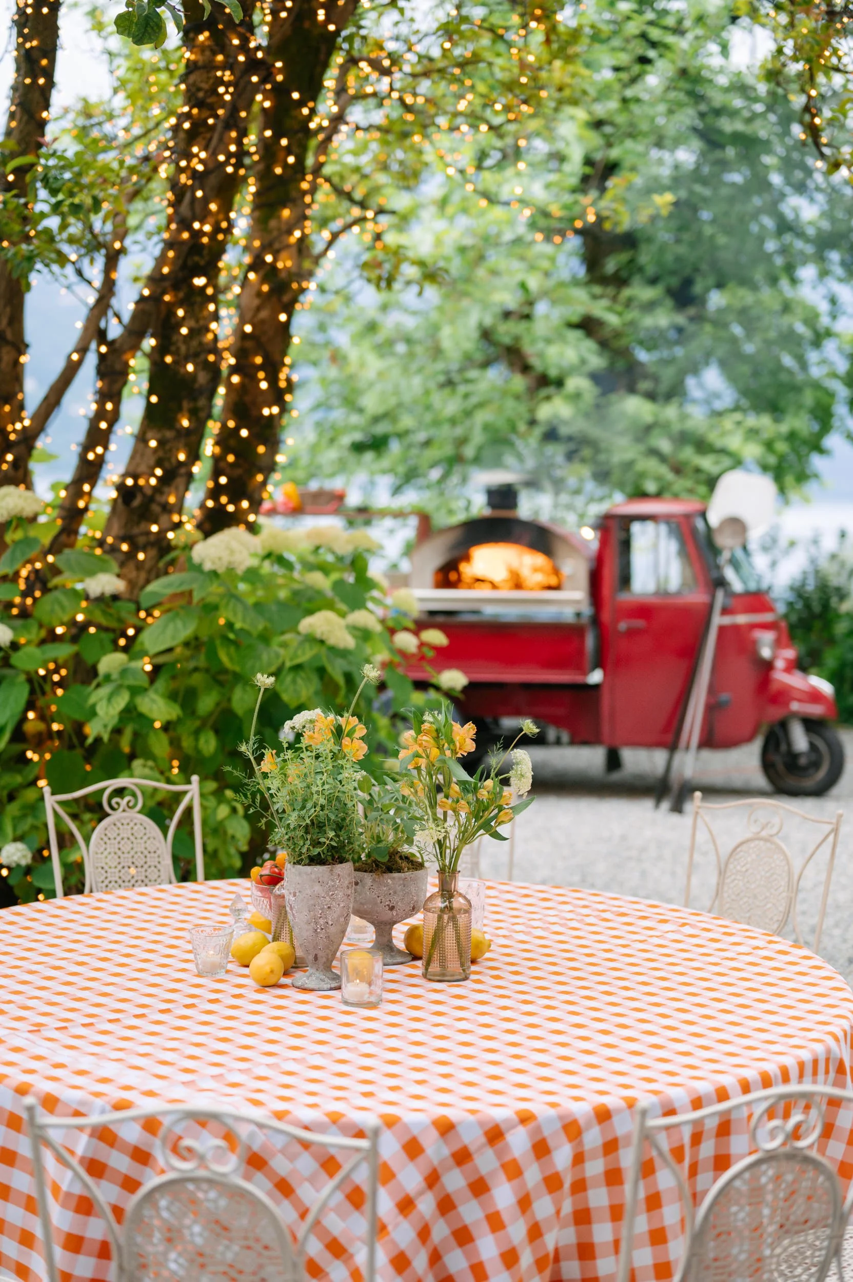 Outdoor dining setup with a round table covered with an orange and white checkered tablecloth, decorated with vases of flowers and lemons, surrounded by white chairs. In the background, a red food truck with an open oven and string lights hanging fro