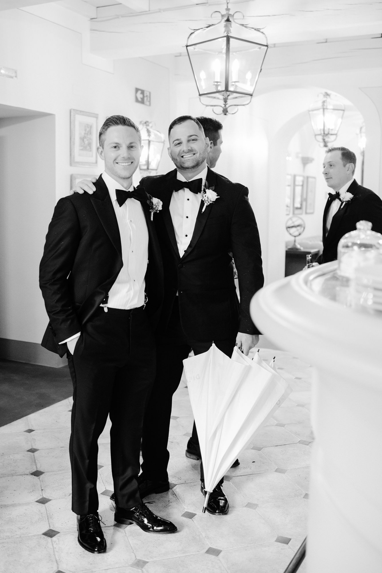 Two men in tuxedos, one holding an umbrella, smiling and posing together indoors.