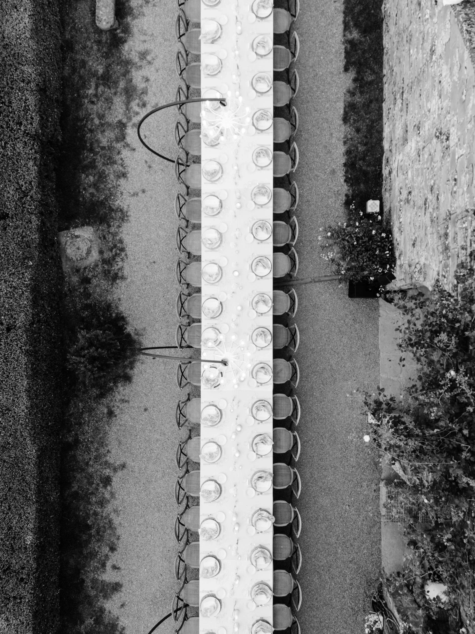Overhead black-and-white photo of a long outdoor dining table set with plates, glasses, and candles, surrounded by chairs. The table is positioned on a gravel surface with bushes and plants on either side.