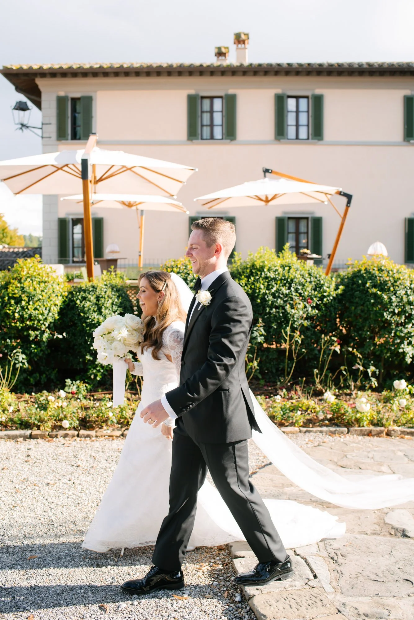 A newlywed couple walking outdoors on a sunny day, with the bride holding a bouquet of white flowers and the groom in a black tuxedo, in front of a house with green shutters and umbrellas.