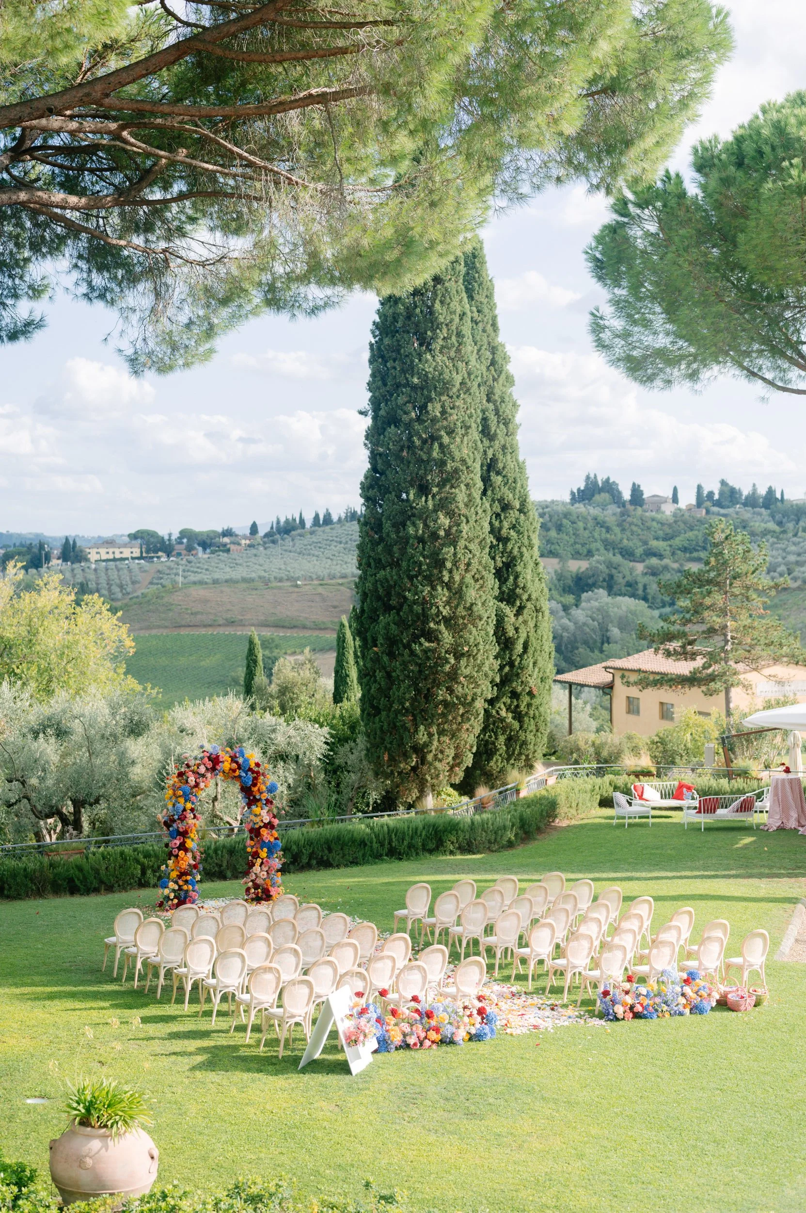 Outdoor wedding setup with chairs arranged in front of a floral arch on a grassy lawn, surrounded by tall trees and scenic countryside.