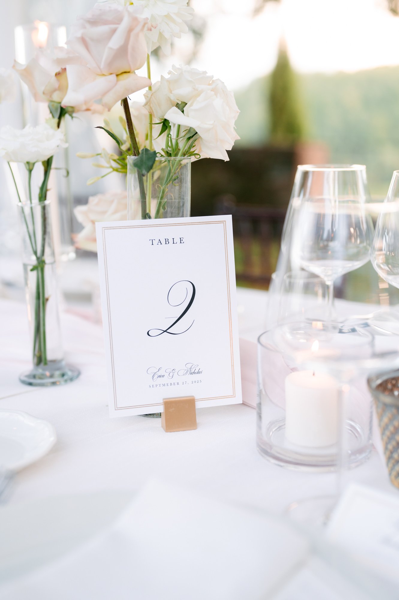 Elegant wedding reception table decor with floral centerpieces, wine glasses, and a table number card for table 2, featuring white and soft pink flowers and candlelight.
