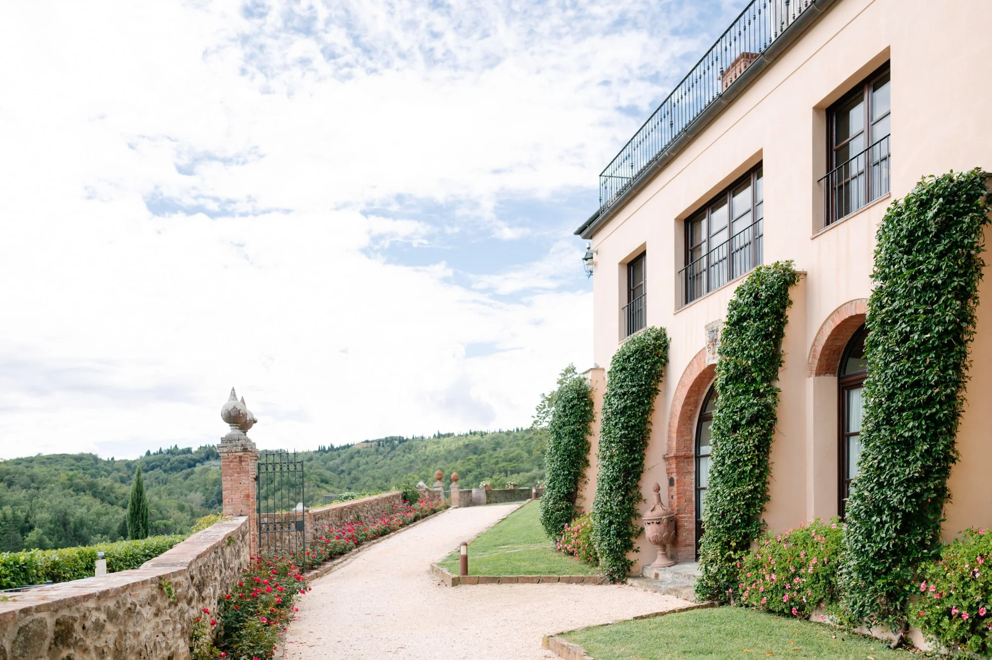 A building with arched windows and green vines growing on the wall, overlooking a scenic landscape with a stone wall, a gravel pathway, and lush greenery.