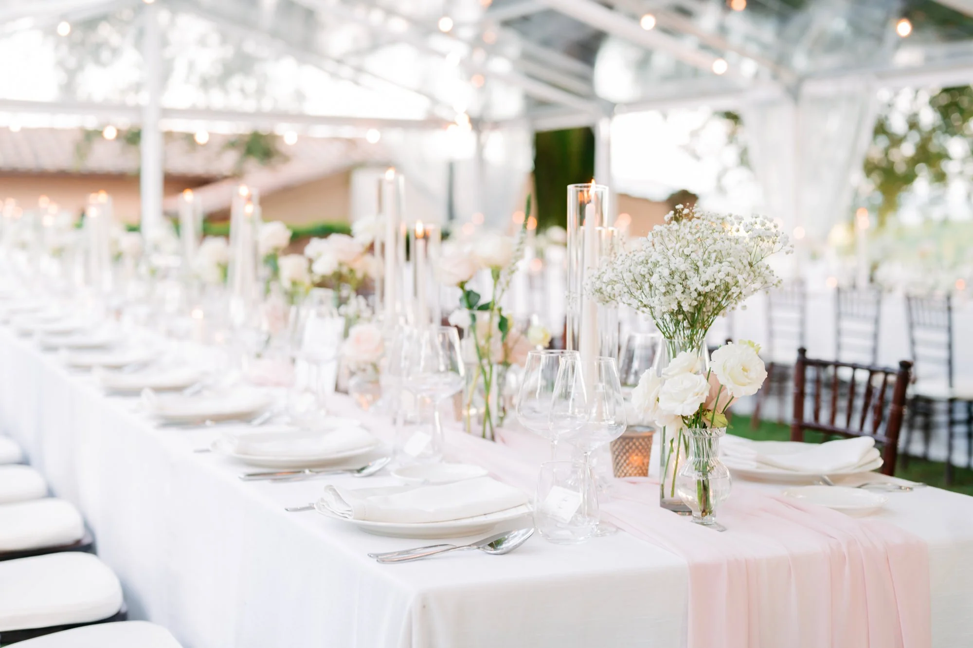 Elegant outdoor wedding reception table decorated with white flowers, candles, and fine glassware under a glass canopy.