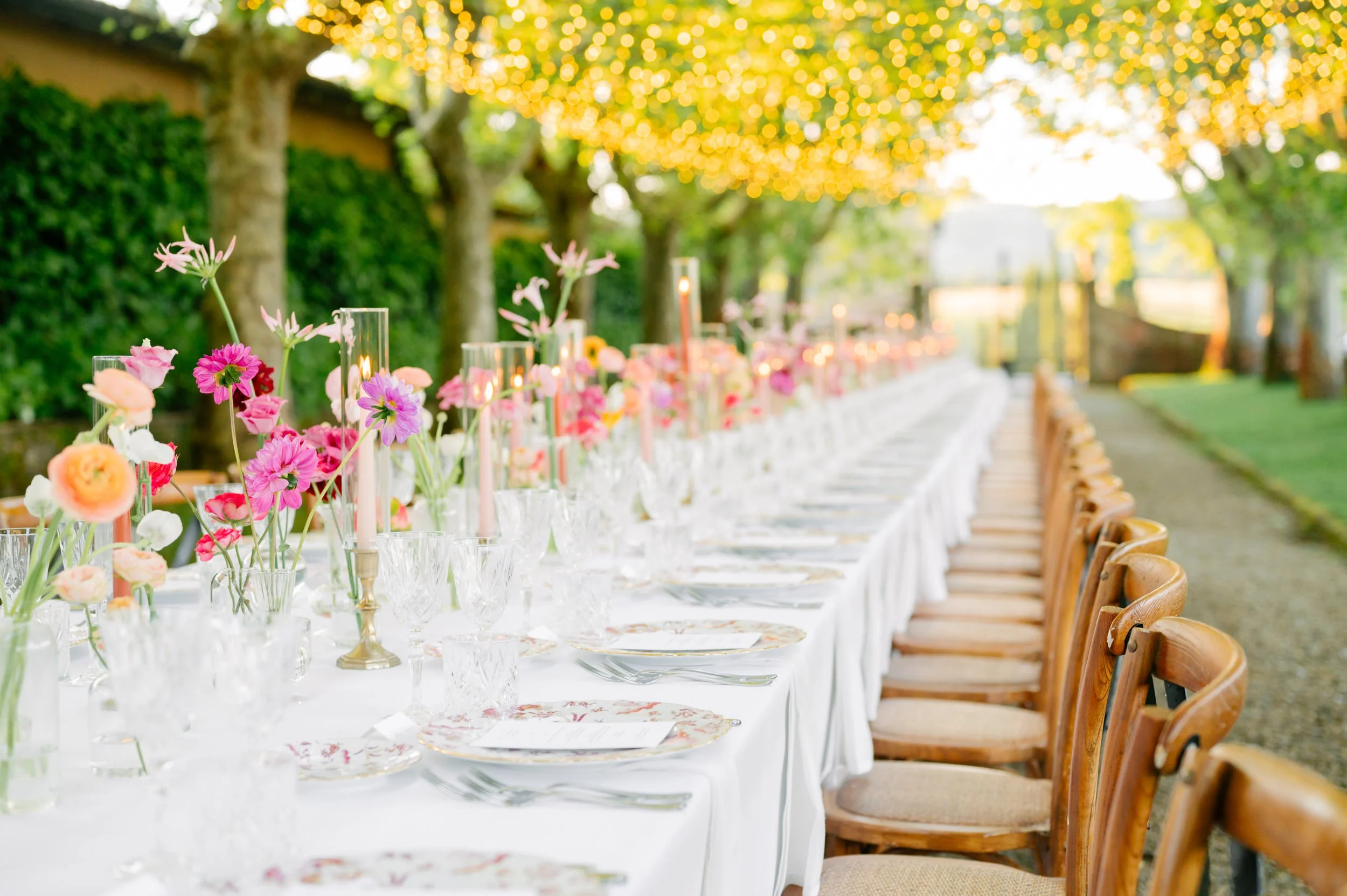 A long outdoor dining table decorated with pink and purple flowers, candles, and glassware, set under string lights in a garden setting.