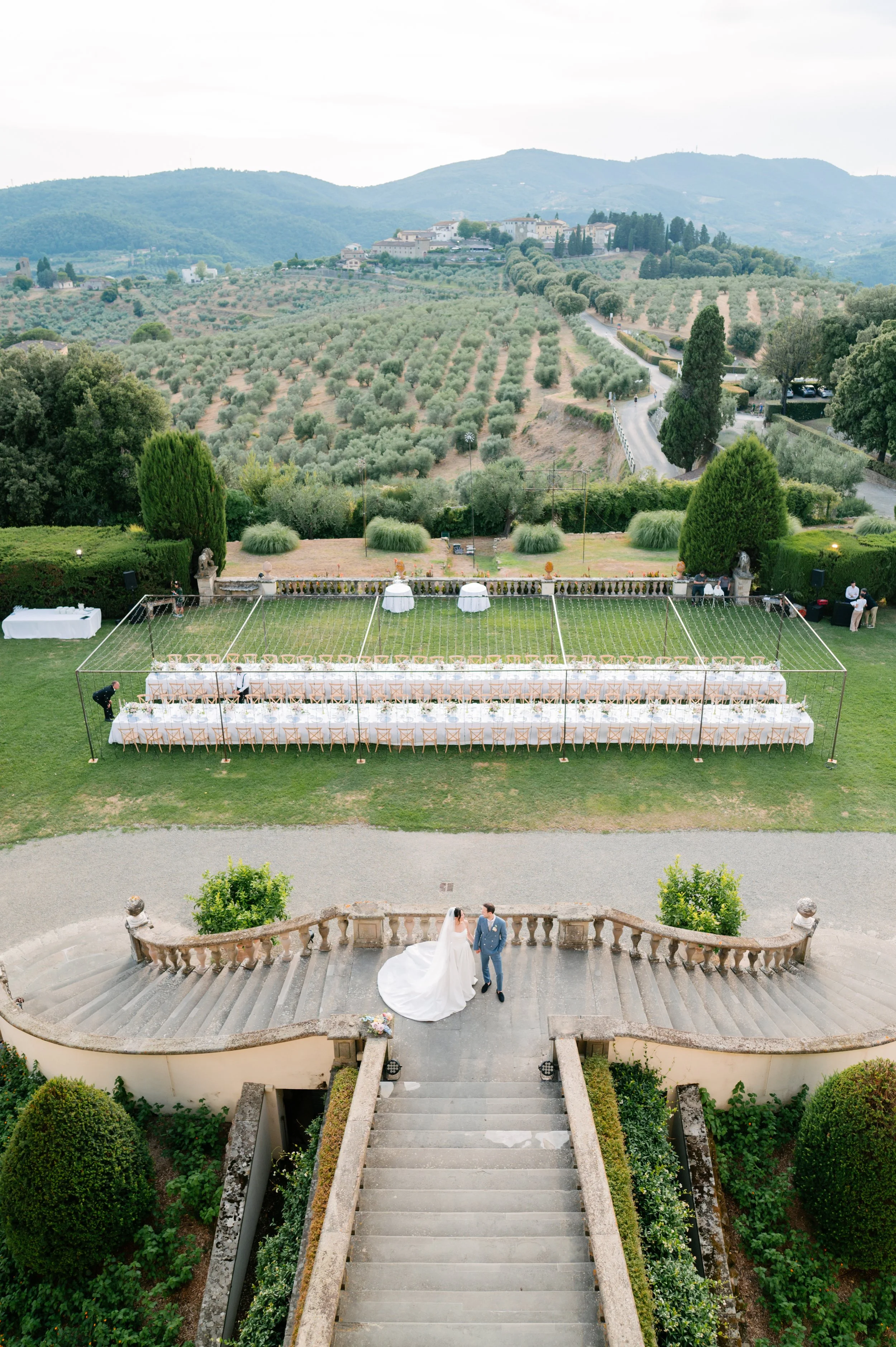 A wedding ceremony setup on an outdoor terrace overlooking rolling hills and a vineyard in the background. A bride and groom stand on the stairs, with the bride in a white wedding gown and the groom in a blue suit. The setup includes a long, decorate