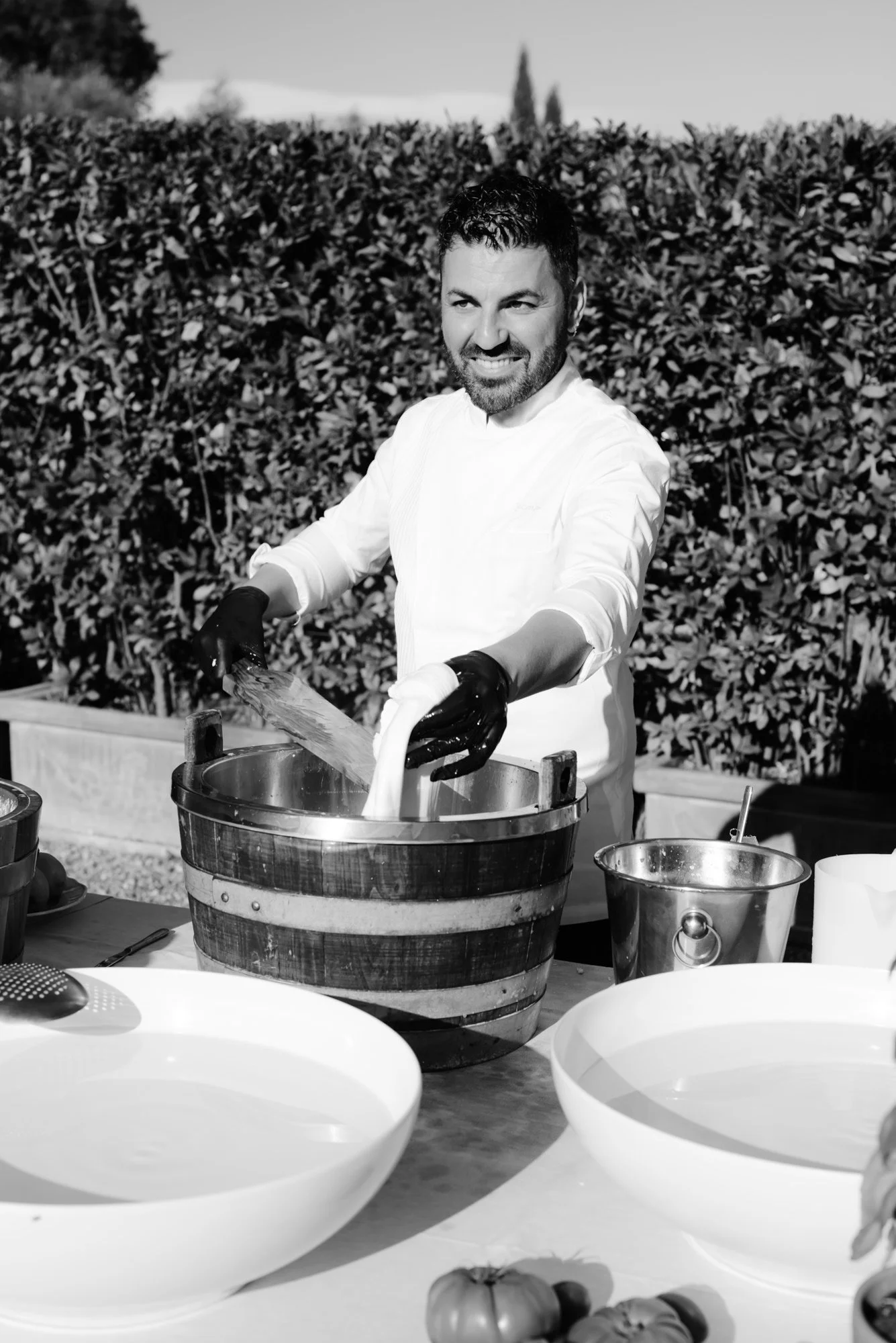 Man in white chef coat and black gloves preparing food outdoors, mixing ingredients in a barrel, with bowls and vegetables on table, hedge background.