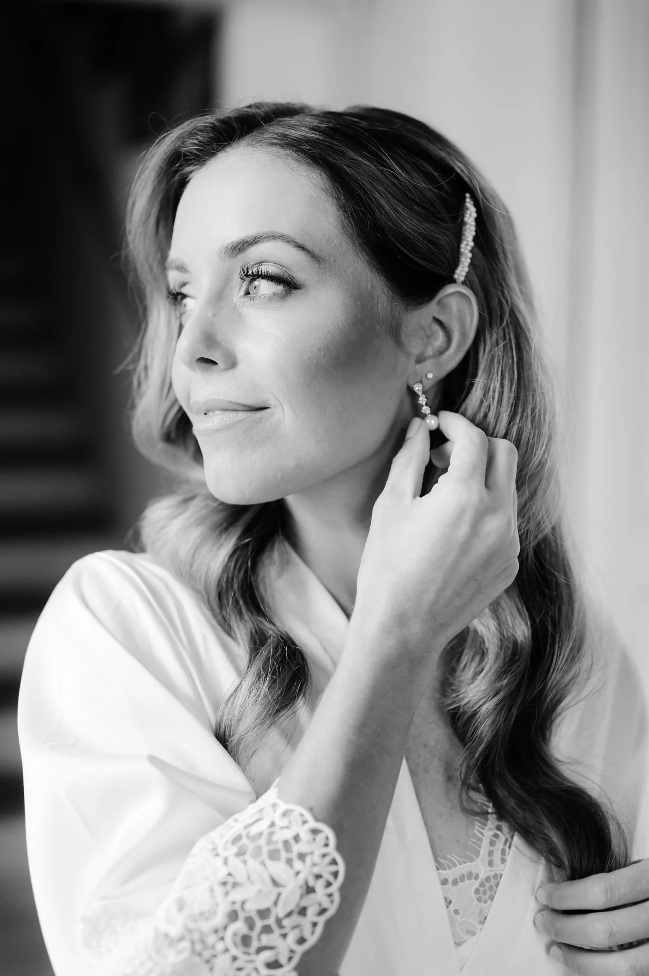 Black and white photo of a woman with wavy hair wearing earrings, fixing her earring with her right hand, smiling gently, dressed in a silk robe with lace detail.