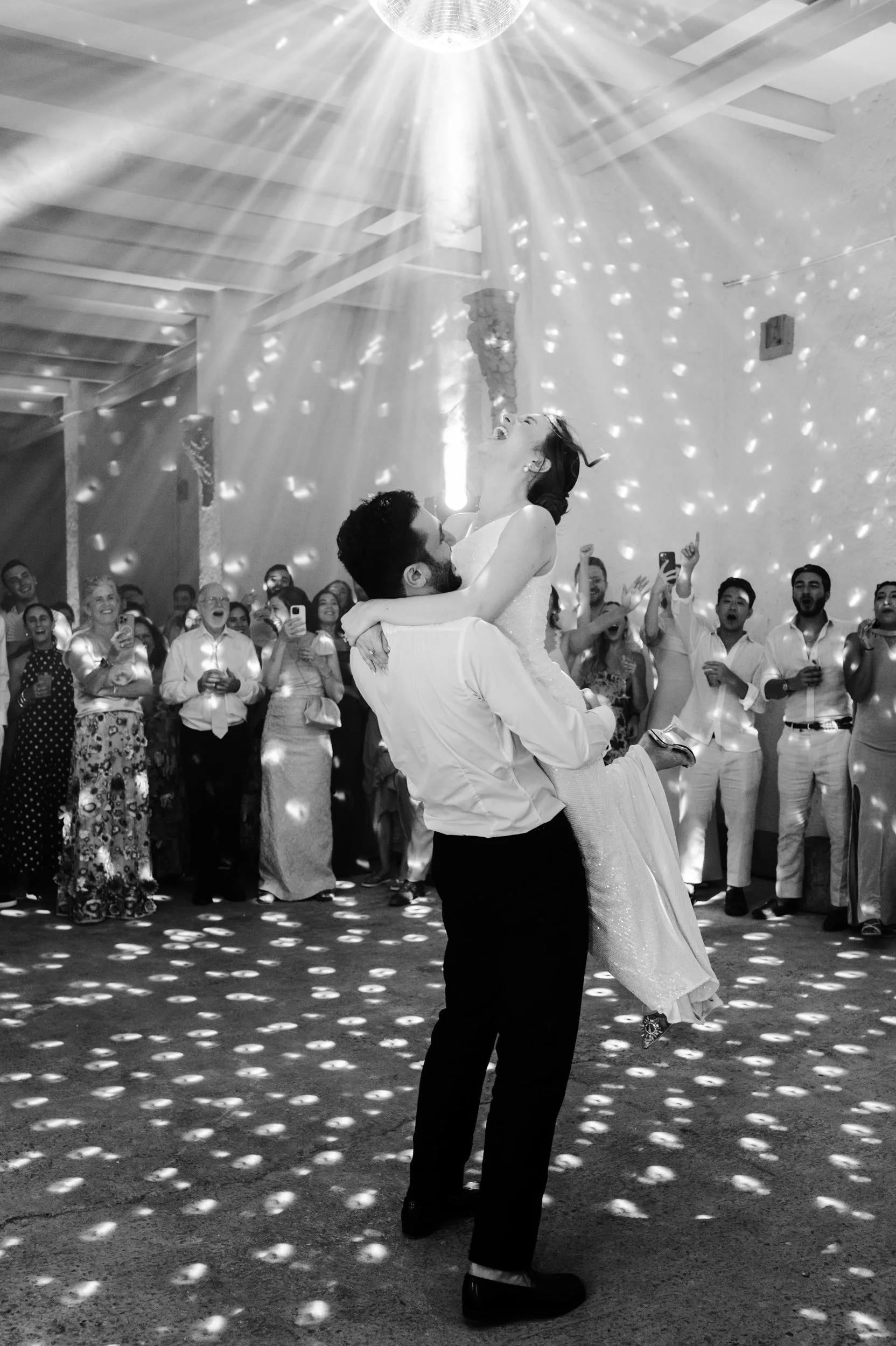 A wedding reception with a couple dancing, the man lifting the woman, surrounded by guests taking photos and cheering, with disco lights creating patterns on the floor and ceiling.