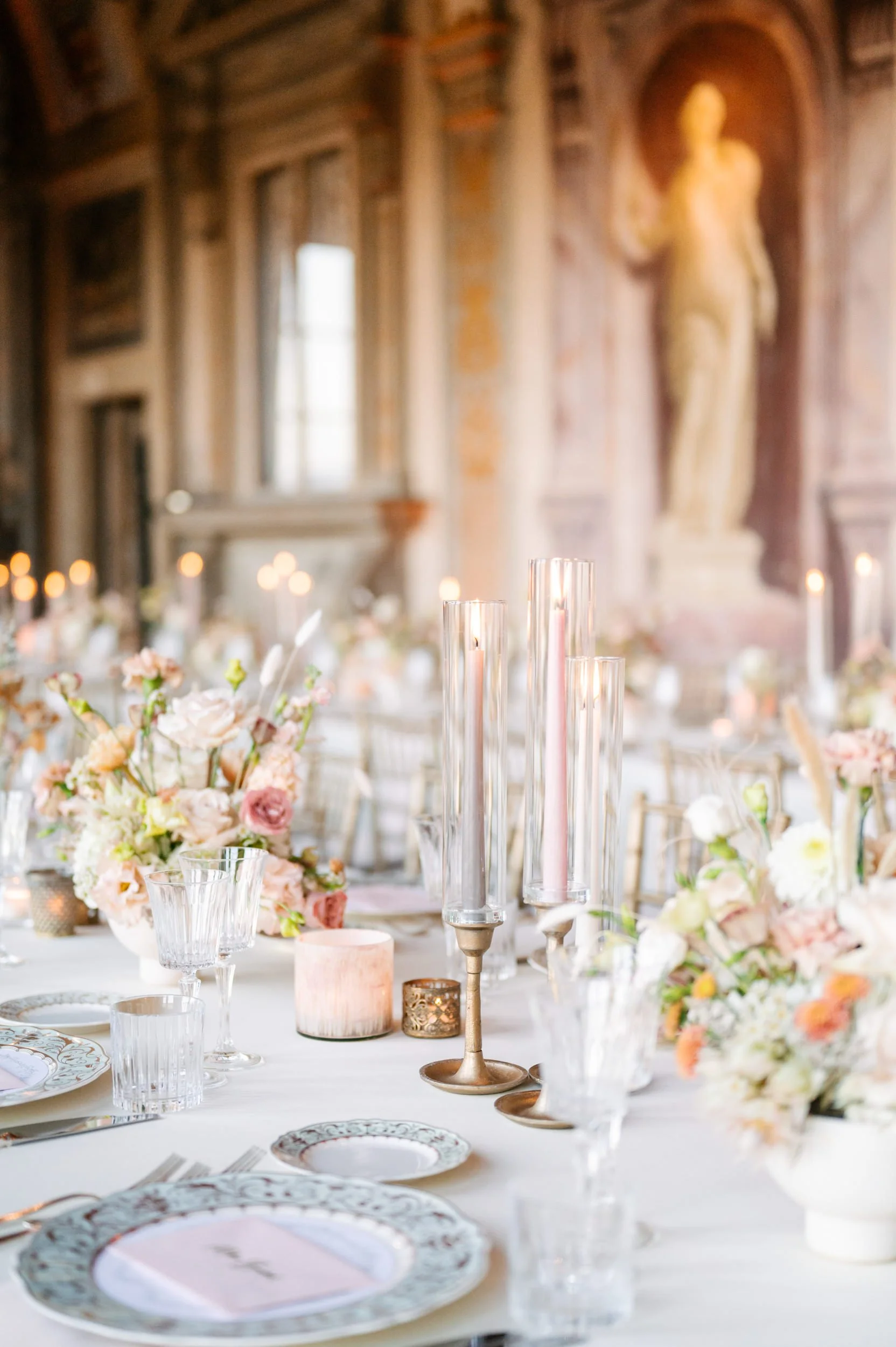 Elegant table setting with pink and white flowers, tall pink candles in glass holders, and ornate plates in a decorated room with a statue of a woman on the wall.