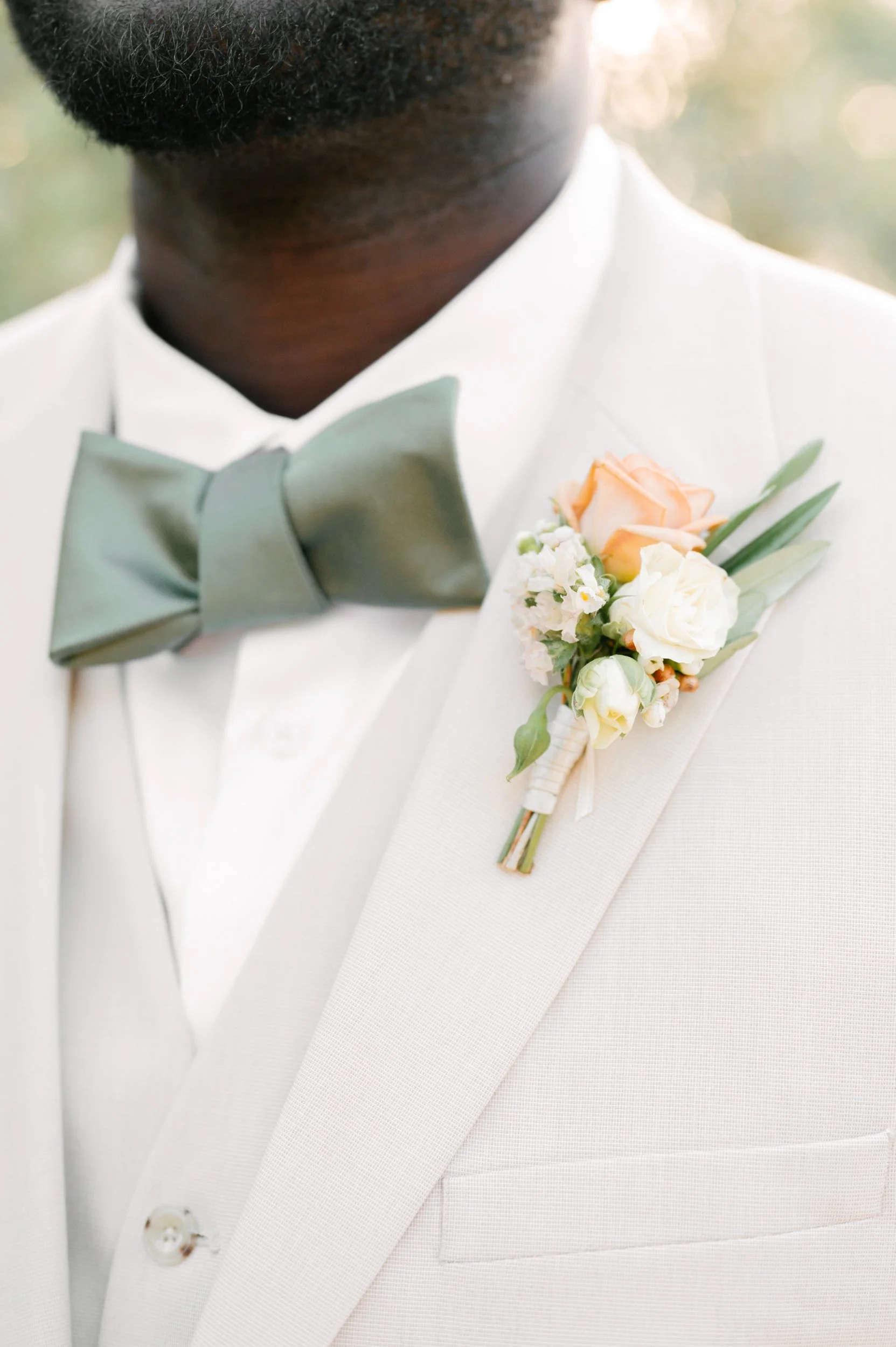 Close-up of a groom in a white tuxedo with a light gray bow tie and a floral boutonniere, featuring roses and greenery.