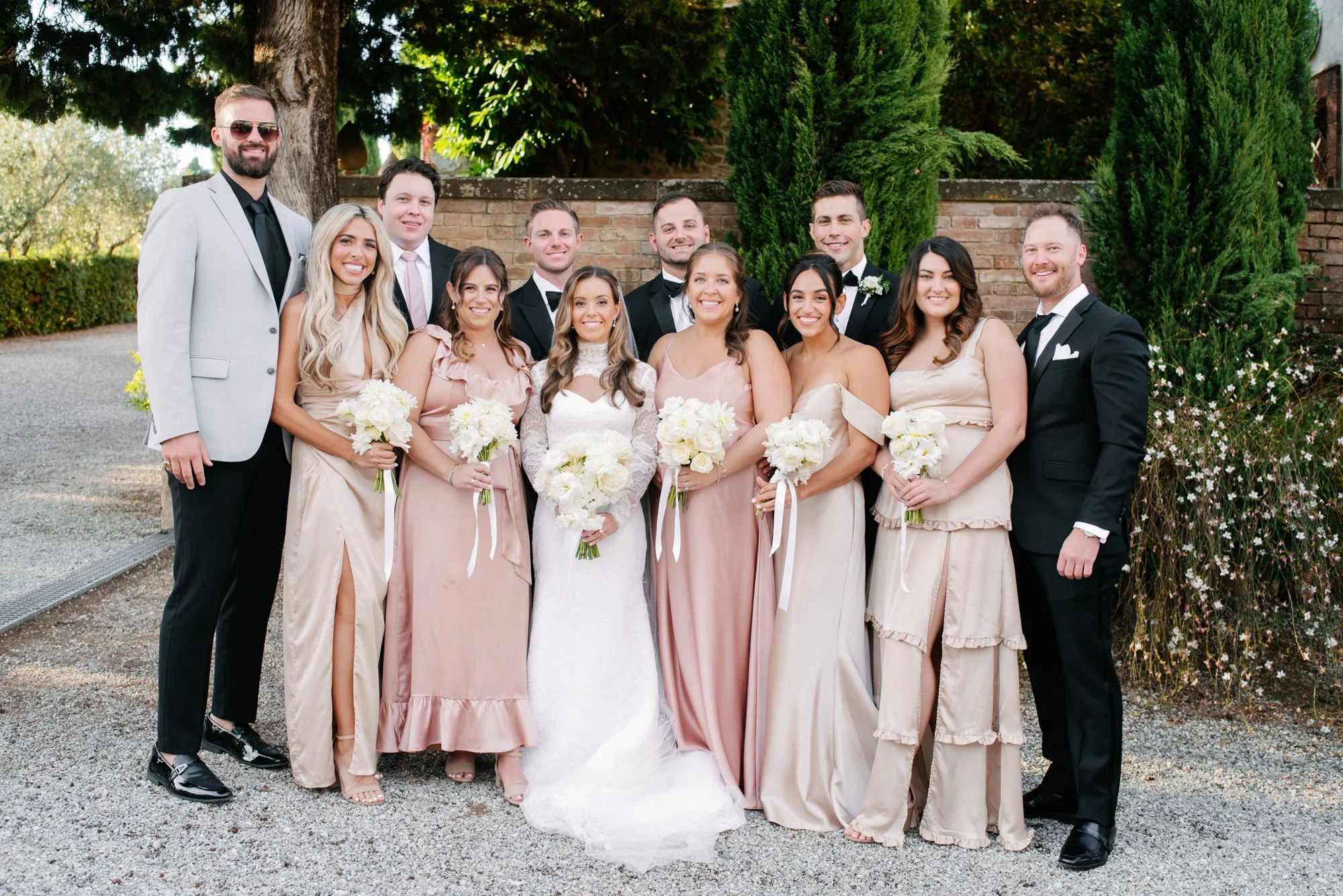 A wedding party of 12 people, including the bride in a white wedding gown holding a white bouquet, bridesmaids in blush dresses holding white bouquets, and groomsmen in suits and tuxedos, standing outdoors in front of greenery and a brick wall.