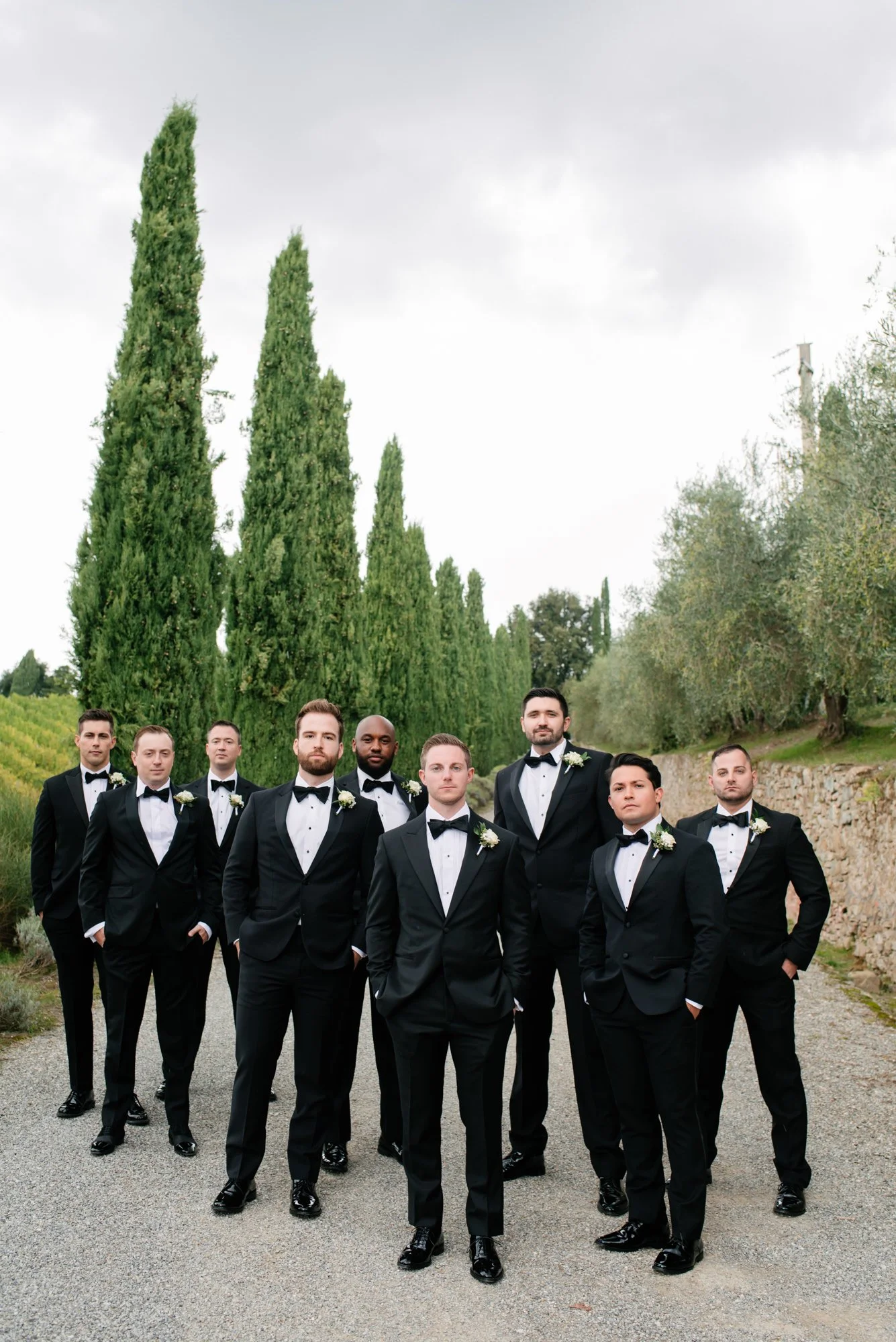 Group of nine men dressed in black tuxedos with bow ties, standing outdoors on a gravel path, with tall cypress trees and a stone wall in the background.
