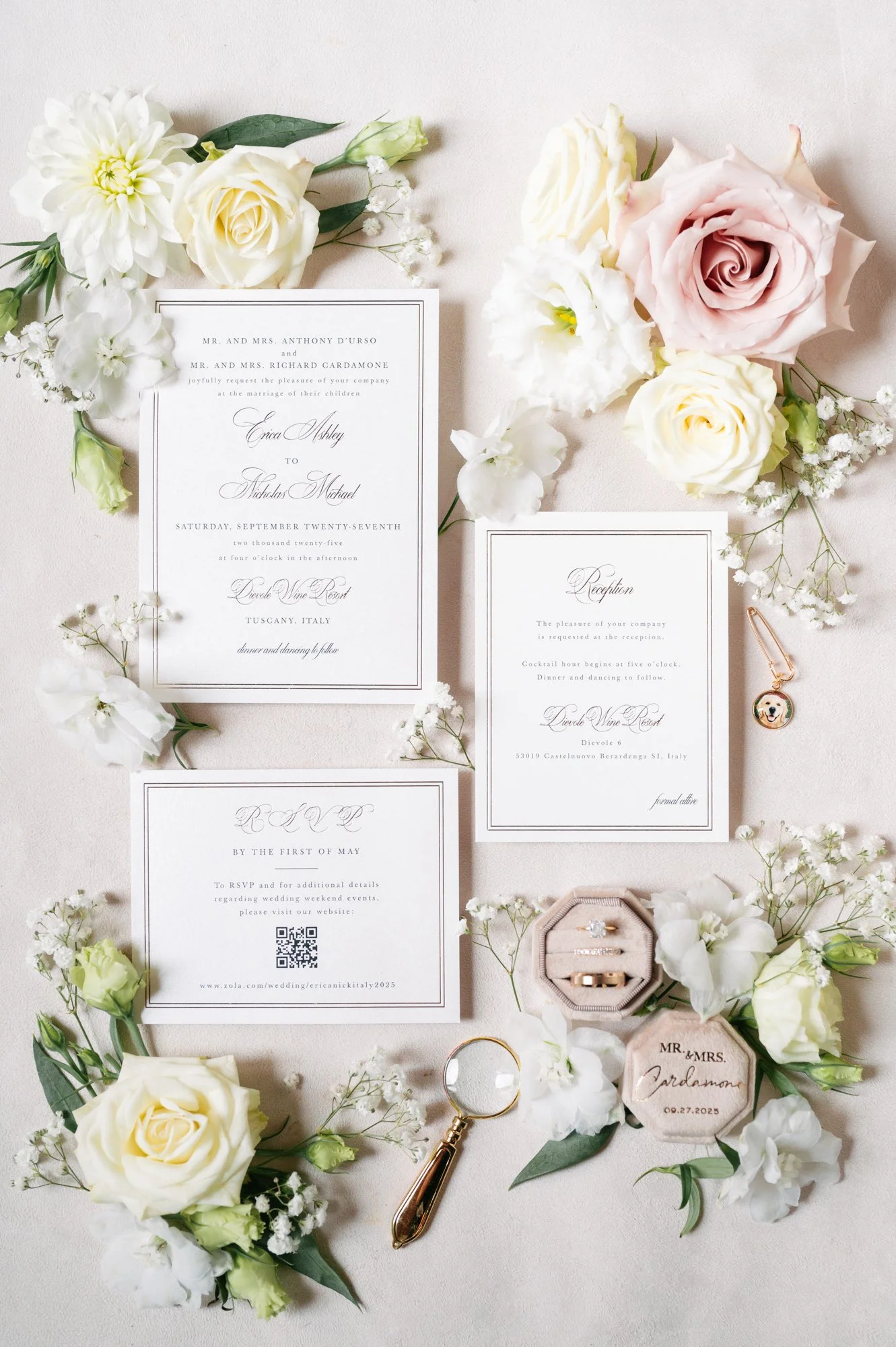 Wedding invitation suite with floral accents, rings, a magnifying glass, and a charm with a dog's face, arranged on a light-colored background.