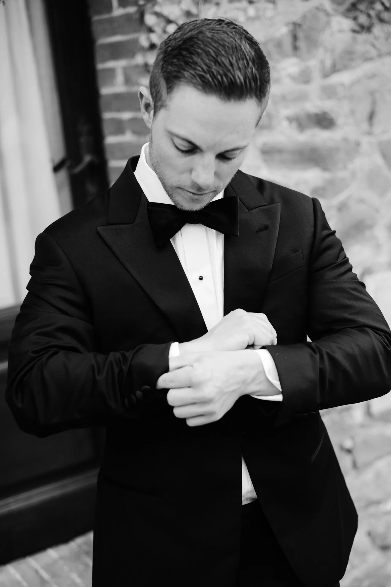 Black and white photo of a man in a tuxedo adjusting his cufflinks, with a brick wall background. Luxury editorial wedding photography in Tuscany. Destination wedding in Italy. L&V Photography Letizia Maccarini.