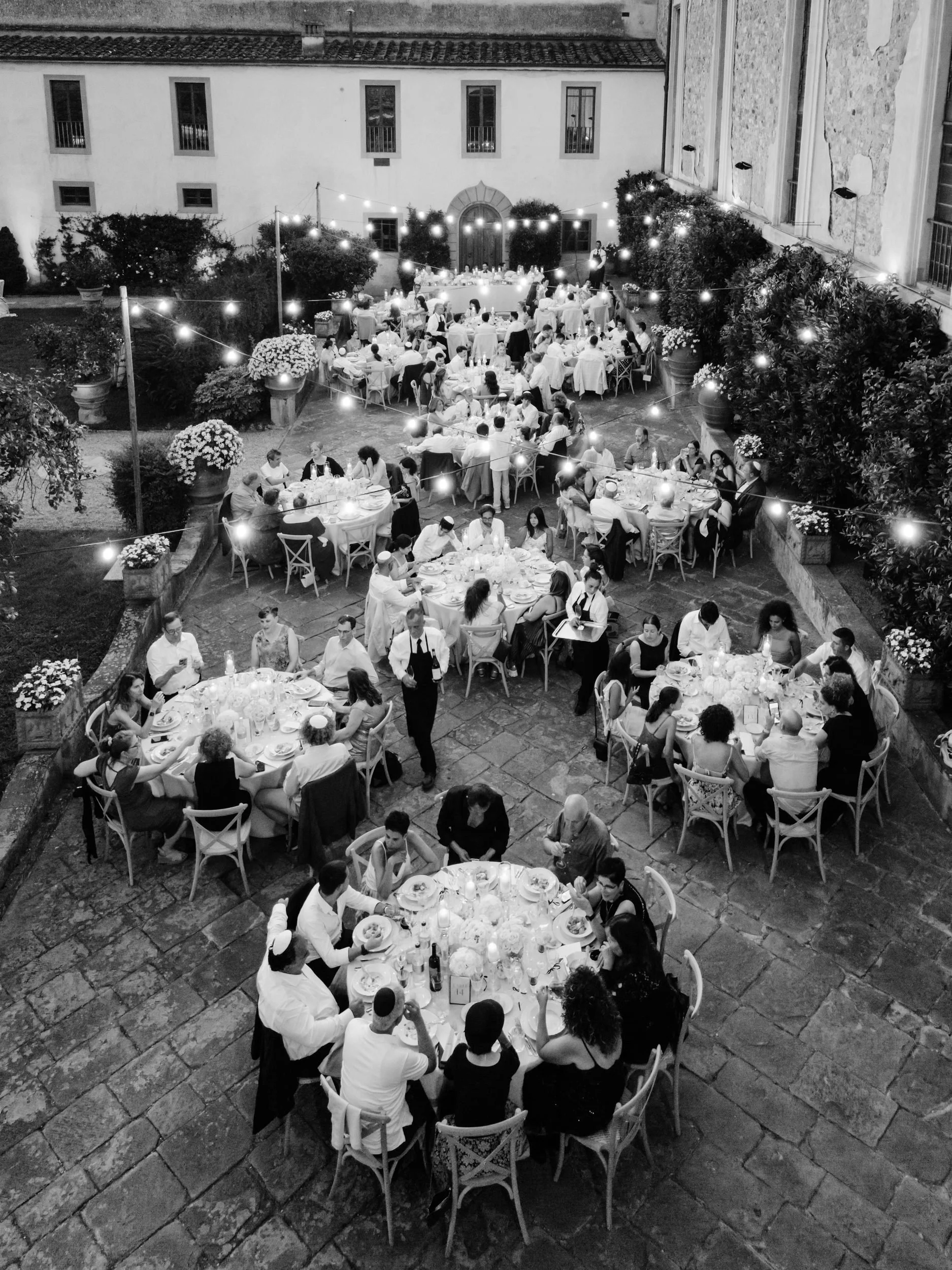 Black and white photo of an outdoor dinner party with multiple round tables, string lights, and people dining, set in a courtyard surrounded by building walls with windows and trees.