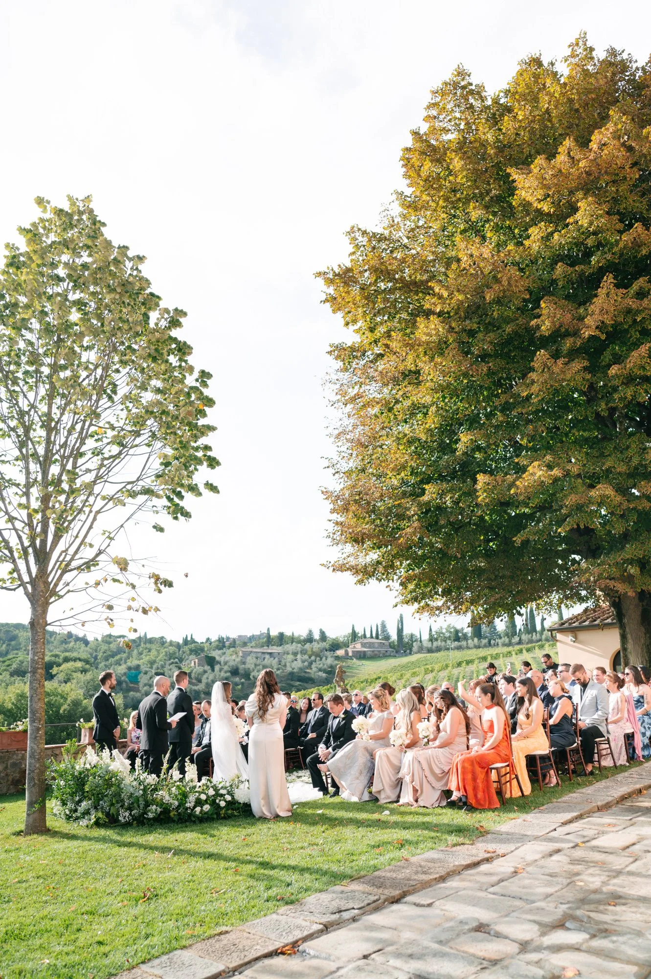 Outdoor wedding ceremony with guests seated on the grass, officiant, bride, and groom under large trees on a sunny day with scenic background.