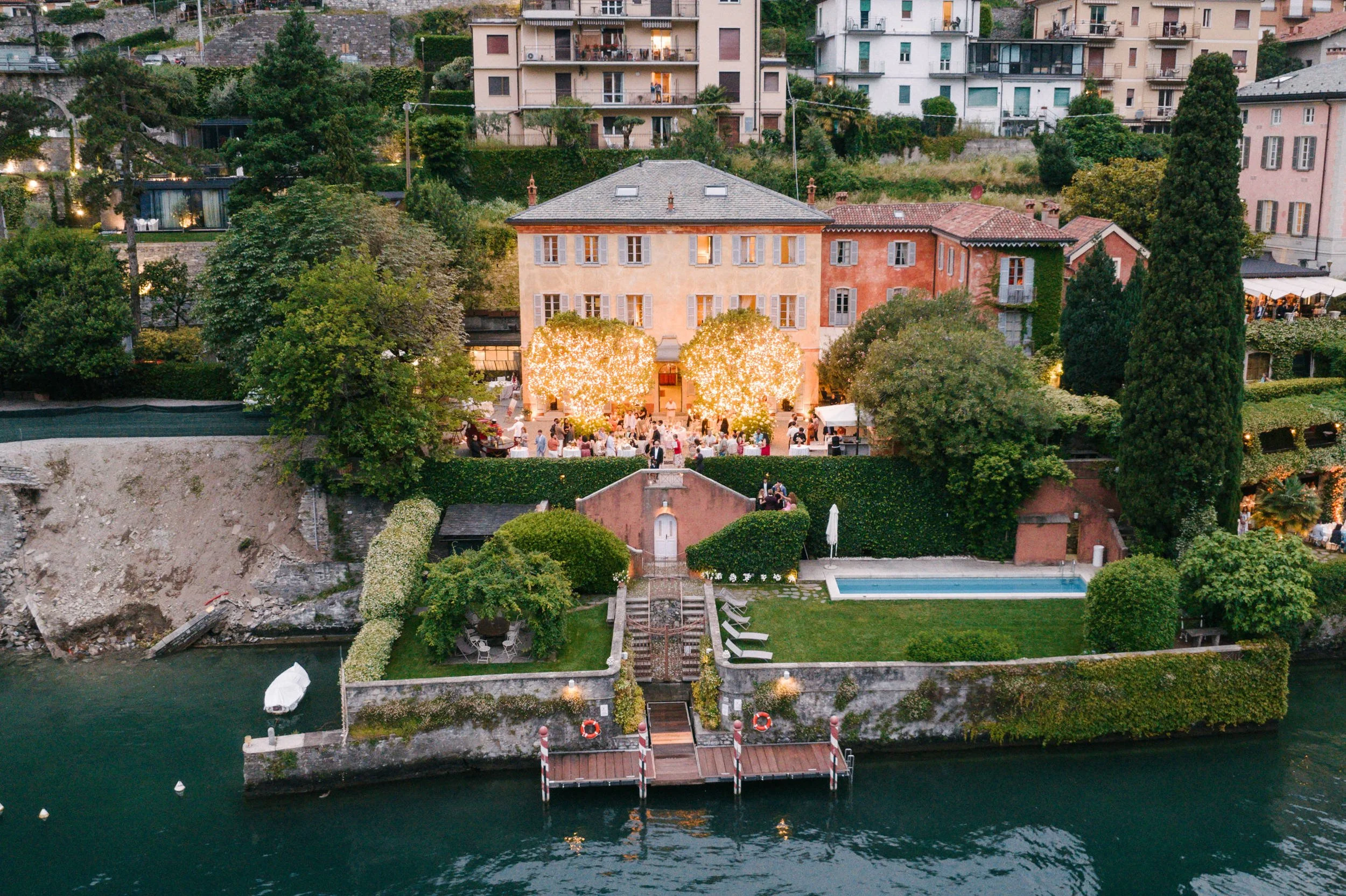 Aerial view of a lakeside event with a yellow building, outdoor party with string lights, green trees, and a swimming pool, surrounded by hillside houses.