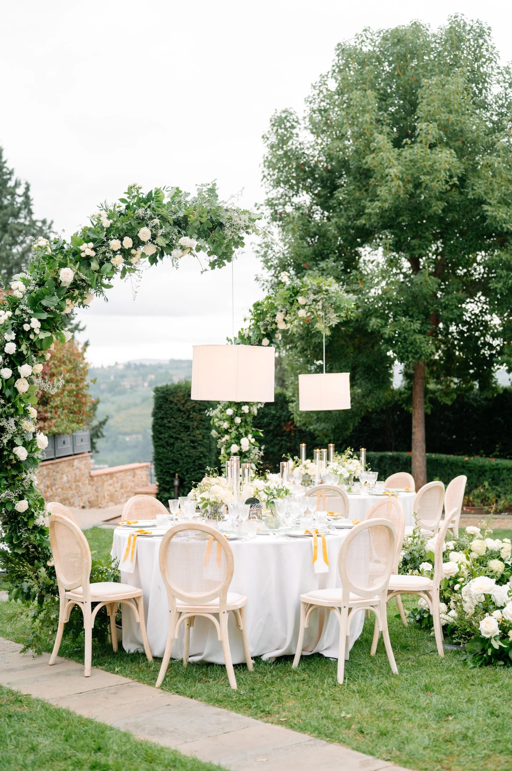 Elegant outdoor wedding reception table set with white linens, floral centerpieces, candles, and surrounded by white chairs, with a large floral arch and lush greenery in the background.