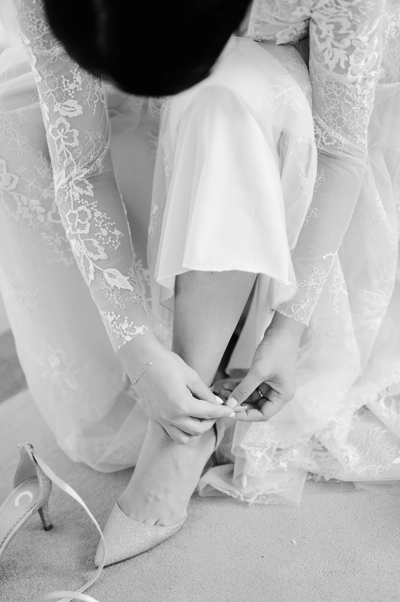 A bride in a lace wedding dress helps a bride with her shoes, focusing on her leg and the act of fastening her shoe strap.