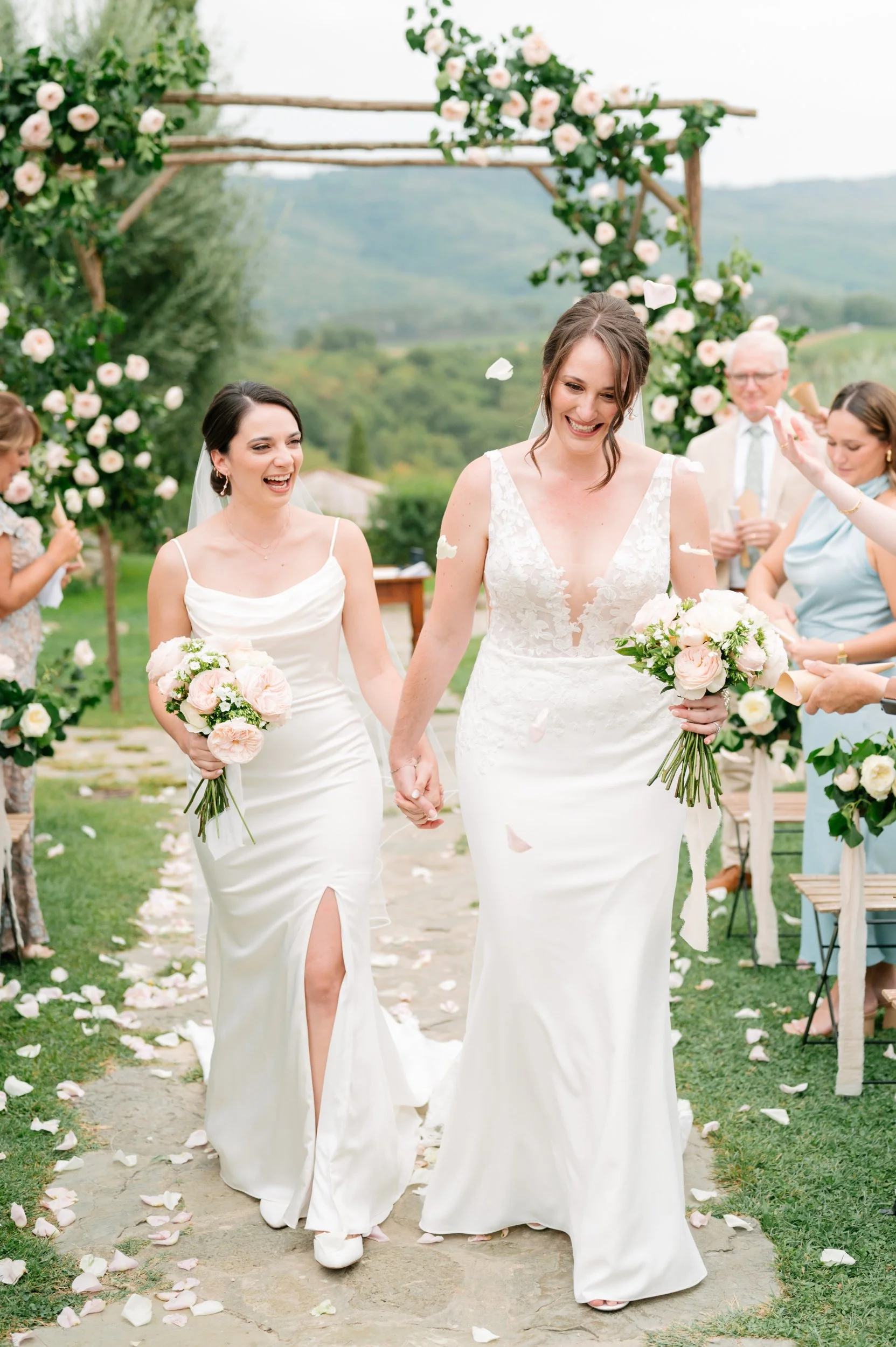 Two brides in wedding dresses holding bouquets, walking hand in hand through a garden aisle, smiling and celebrating at a wedding ceremony.