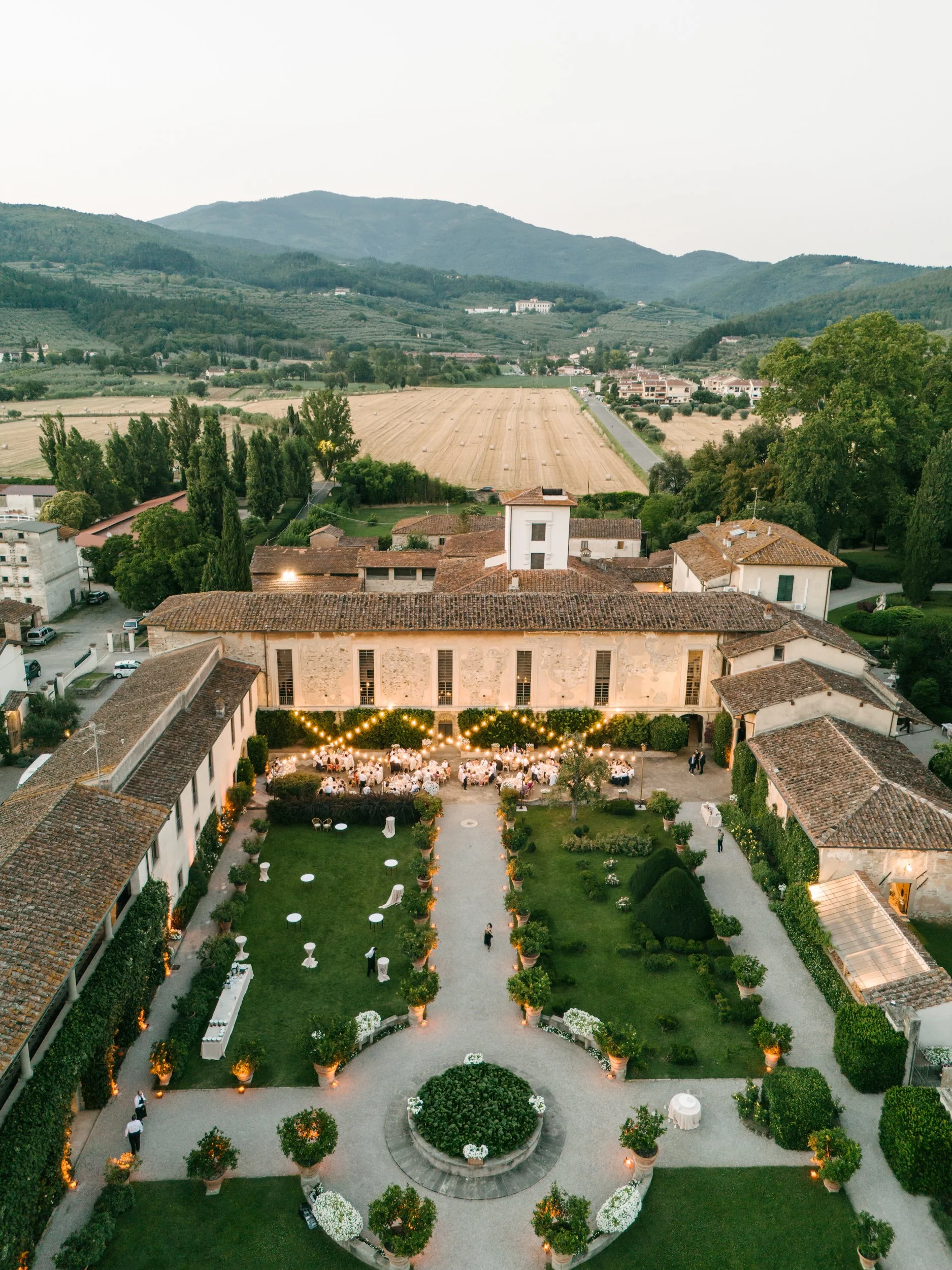 Outdoor wedding reception at an Italian villa with string lights, arranged tables, and lush gardens in a scenic mountainous landscape.