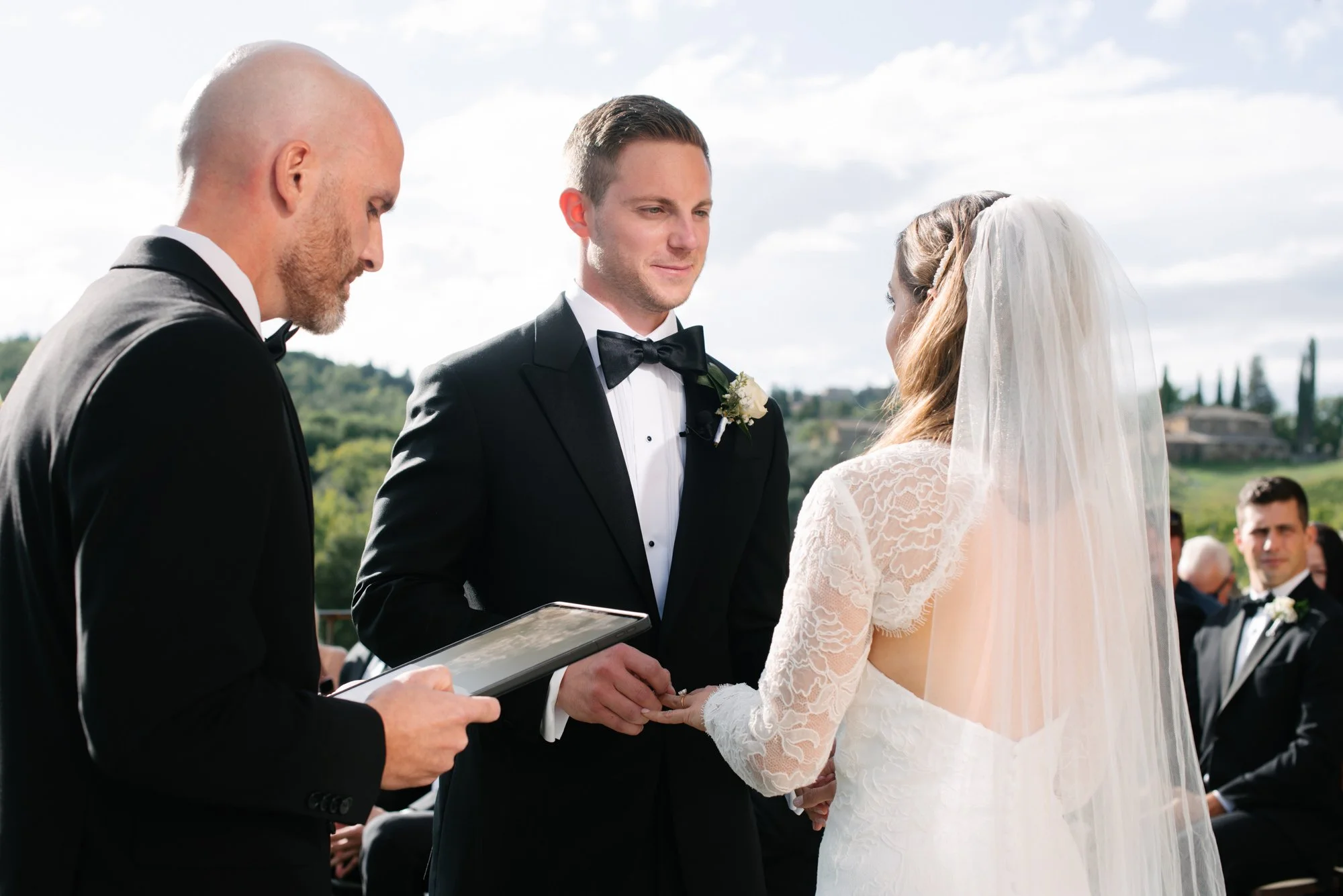 A couple getting married outdoors, with the groom in a black tuxedo and bow tie, holding the bride's hand, and the bride wearing a white lace wedding dress and veil. An officiant stands nearby holding a tablet, officiating the ceremony on a sunny day