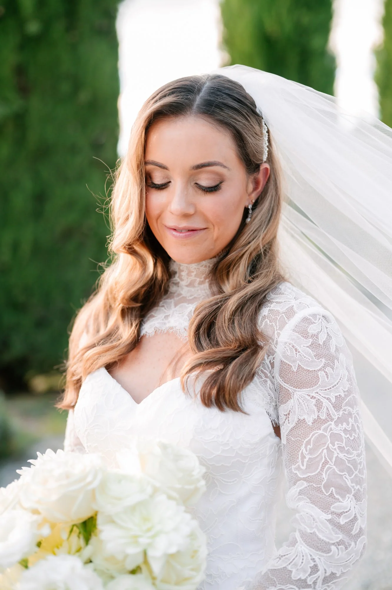 Bride in a lace wedding dress with long wavy hair, holding a bouquet of white flowers, standing outdoors with a blurred green background.