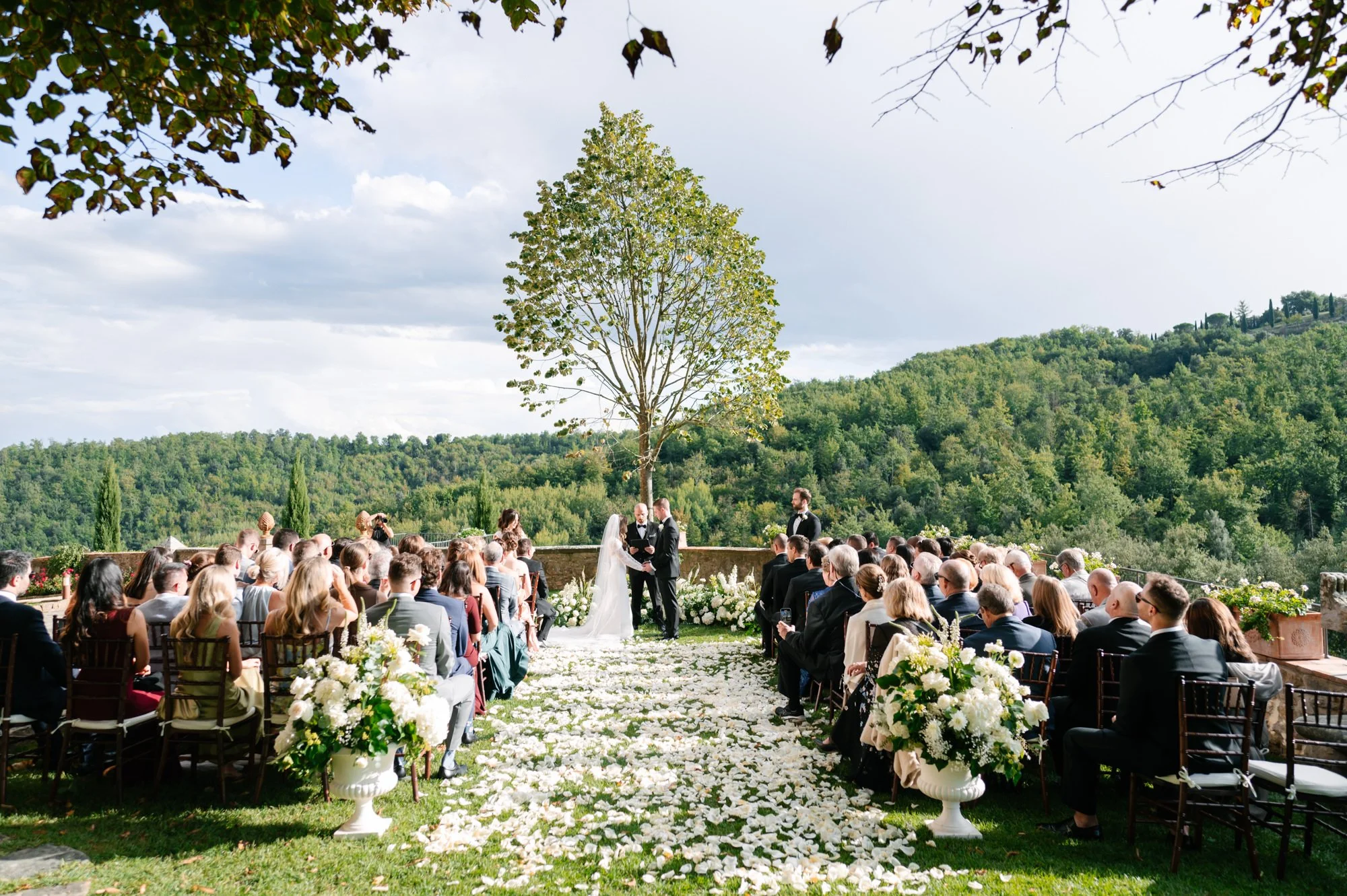 Outdoor wedding ceremony with guests seated on either side of an aisle covered in white flower petals, overlooking a lush green landscape and hill, with a large tree and cloudy sky in the background. Luxury editorial wedding photography in Tuscany. D