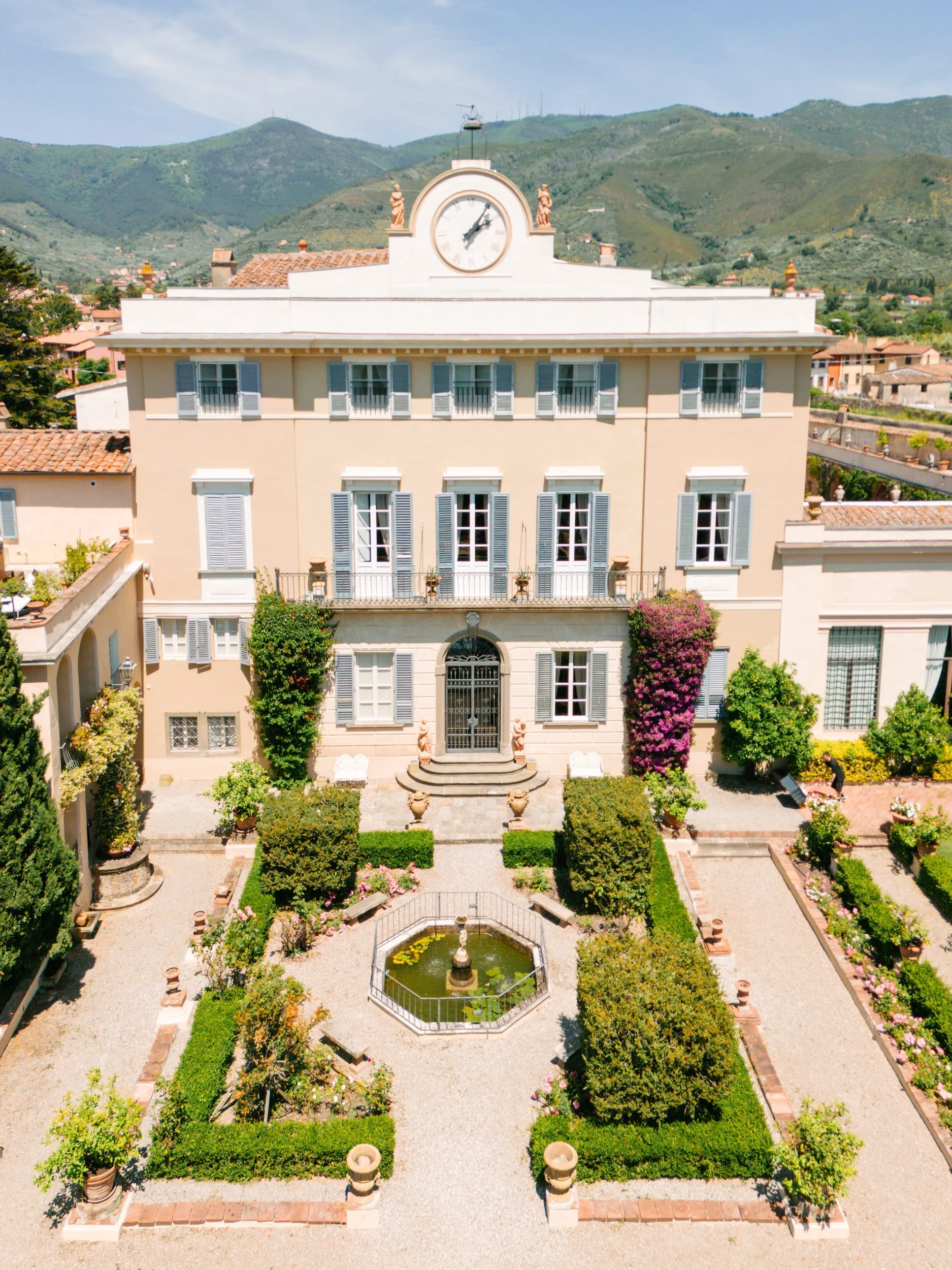 A large, elegant beige mansion with blue shutters, a clock near the roof, and a small fountain in the front yard, surrounded by well-maintained gardens and mountains in the background.