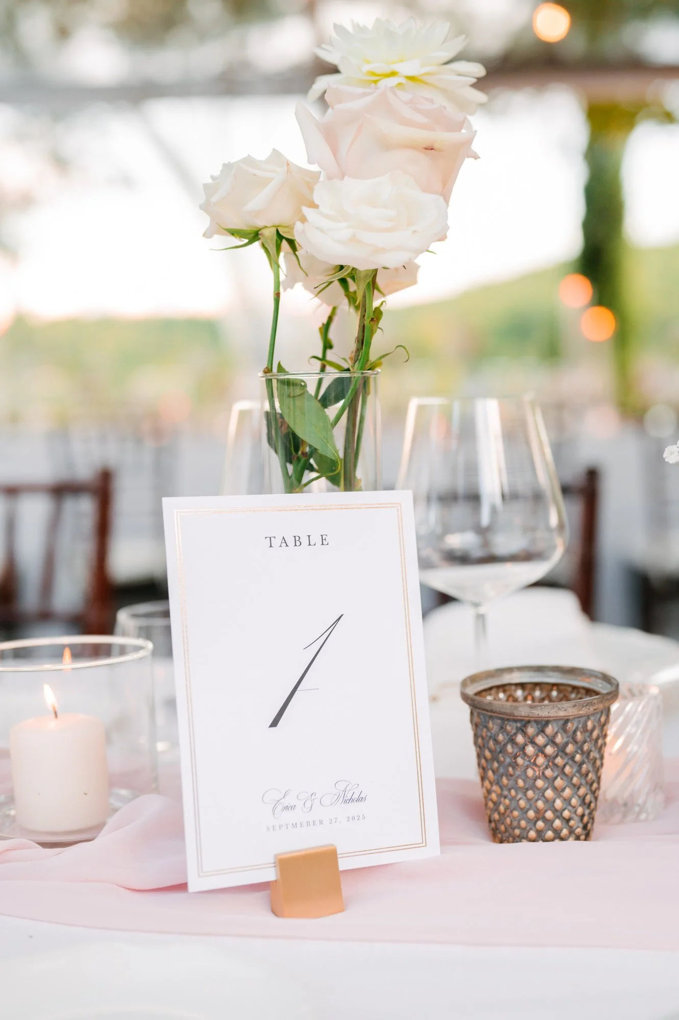 A wedding table centerpiece with a note card, white and blush roses in a glass vase, candles, and decorative candle holders at an outdoor venue with blurred background.
