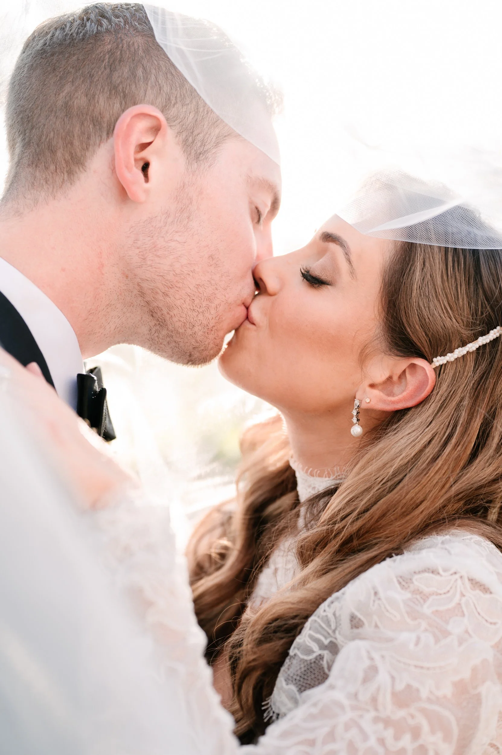 A newlywed couple sharing a kiss, dressed in wedding attire, with the bride wearing pearl earrings and a lace dress, and a veil on her head.