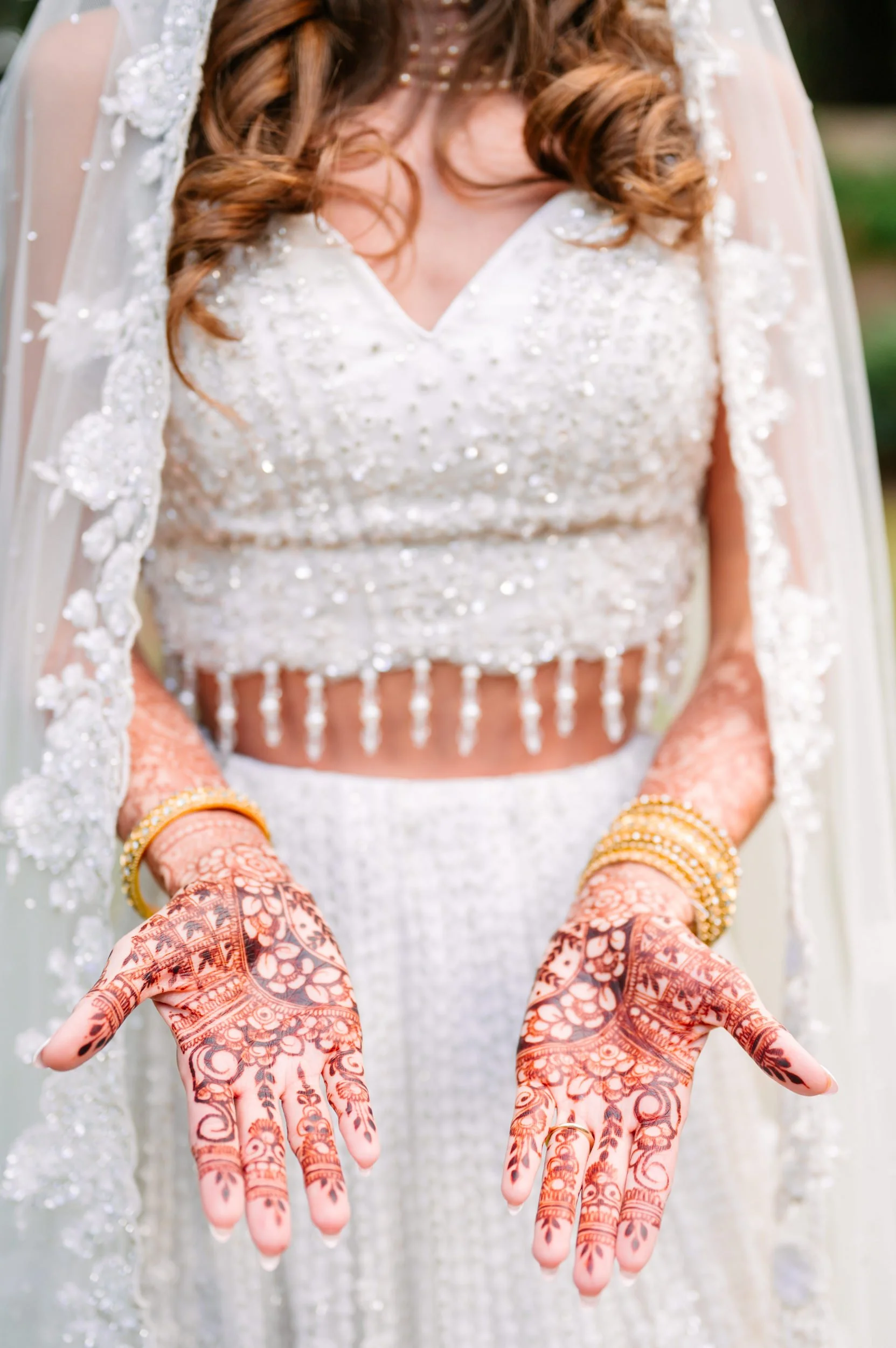 A woman in a white embellished dress displays intricate henna designs on her hands, wearing gold bangles, with a blurred outdoor background.