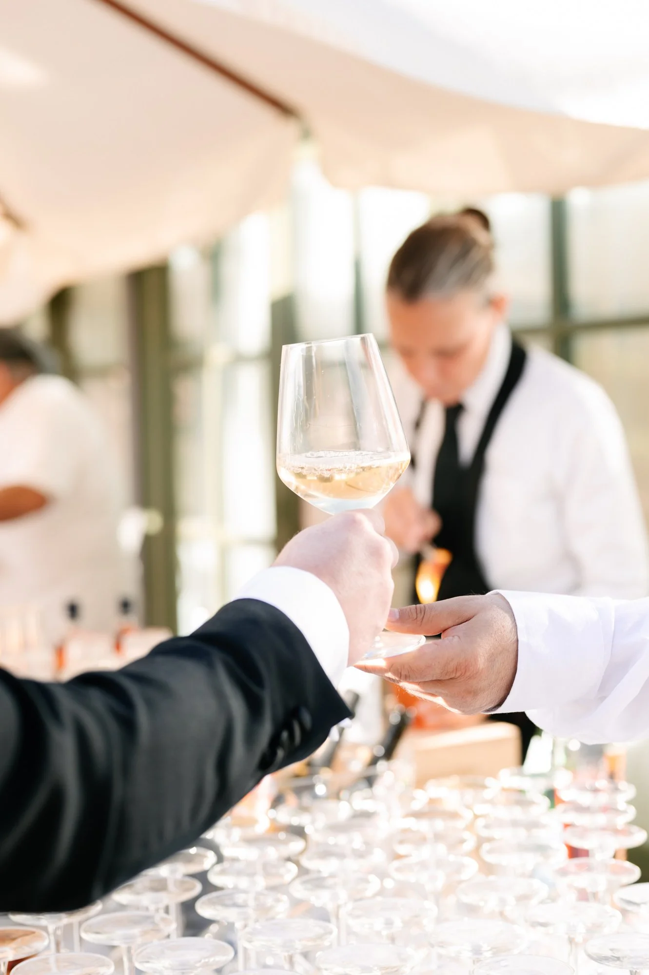 Person in a black suit handing a glass of white wine to another person in a white shirt at a formal event with a bartender in the background.