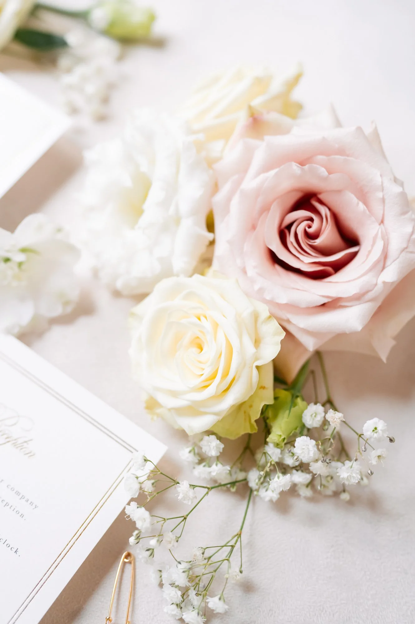 A bouquet of pastel pink, white, and cream roses with white baby's breath flowers on a light-colored surface, next to a partially visible wedding invitation and a gold paperclip.