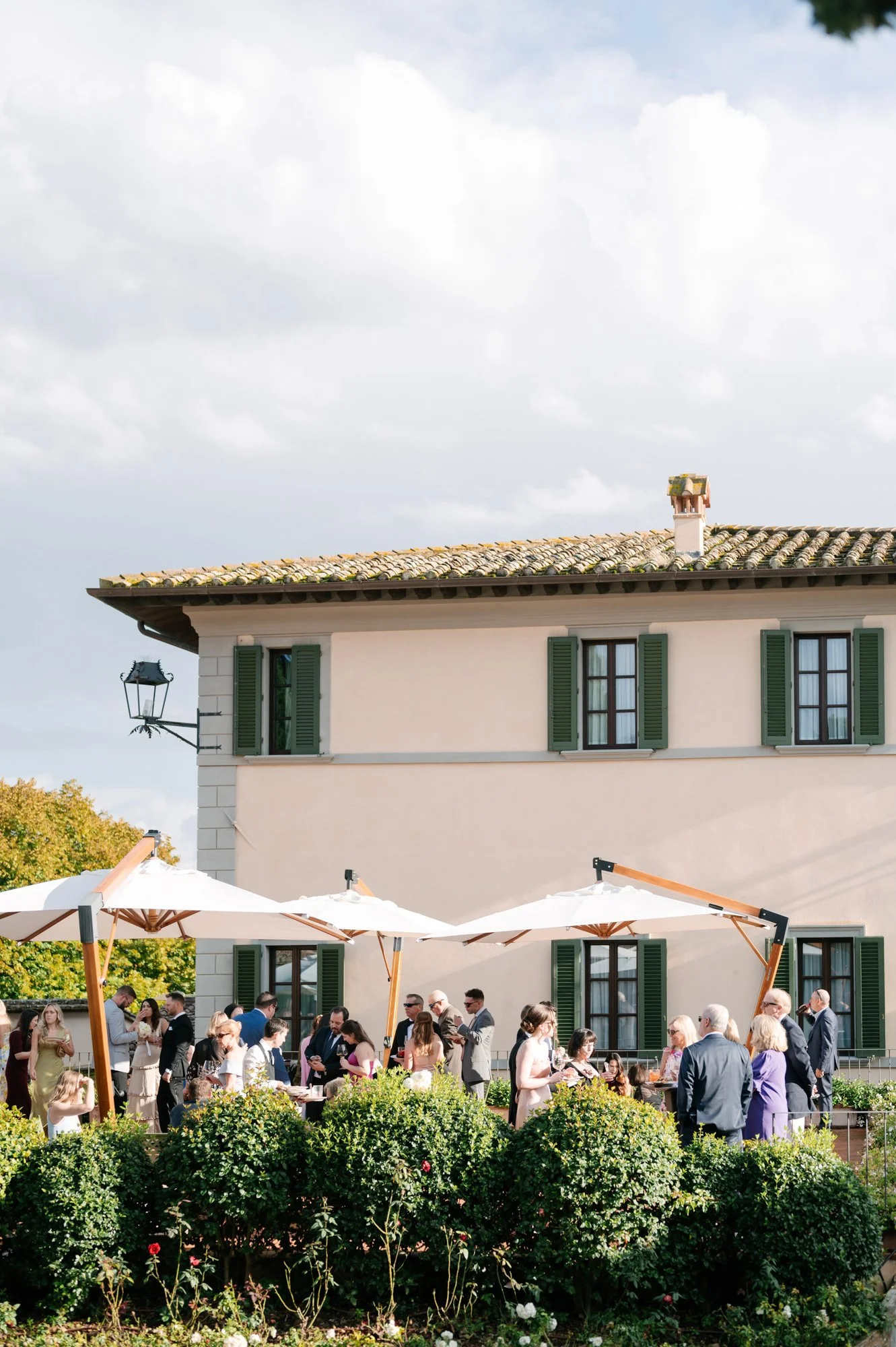 Guests gathered outdoors at a garden party or wedding reception, standing under large white umbrellas in front of a villa-style building with green shutters.