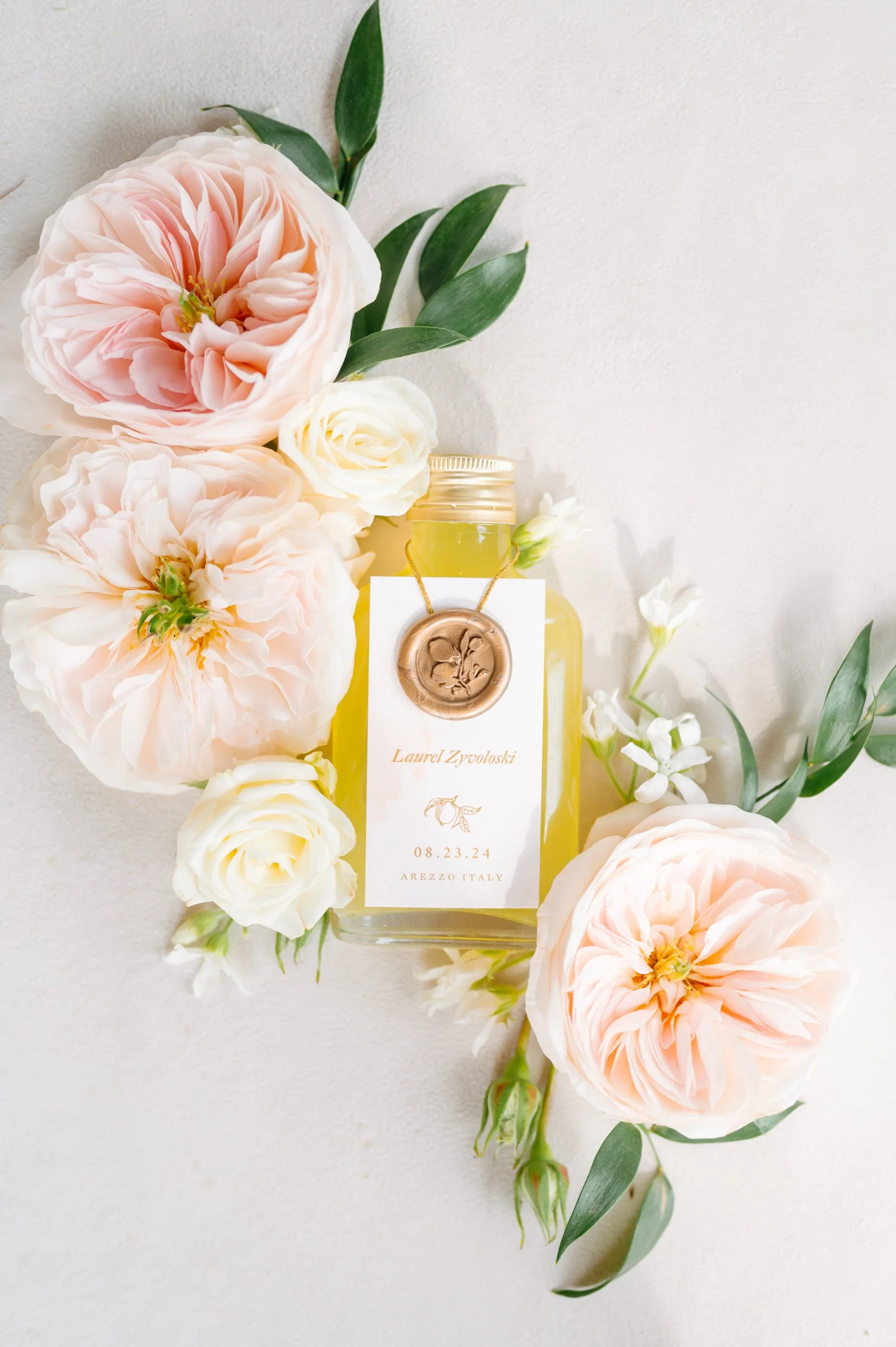 A flat lay of a floral arrangement with pink and white flowers, greenery, and a glass jar with a label. The label has handwritten names, a date, and location, indicating a wedding or special event.