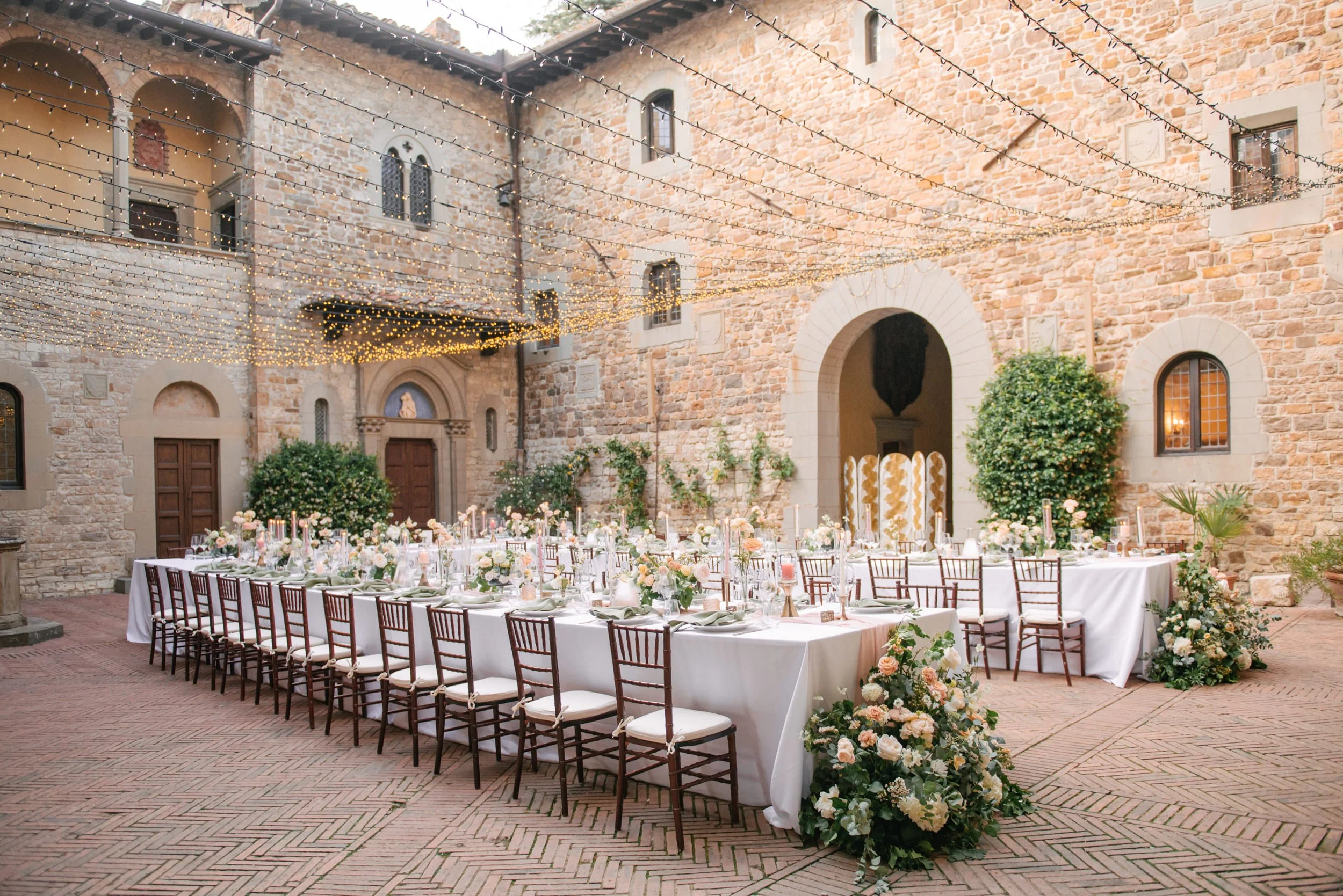 Elegant outdoor wedding reception setup in a historic stone courtyard with string lights overhead, long tables with white tablecloths, and floral centerpieces.