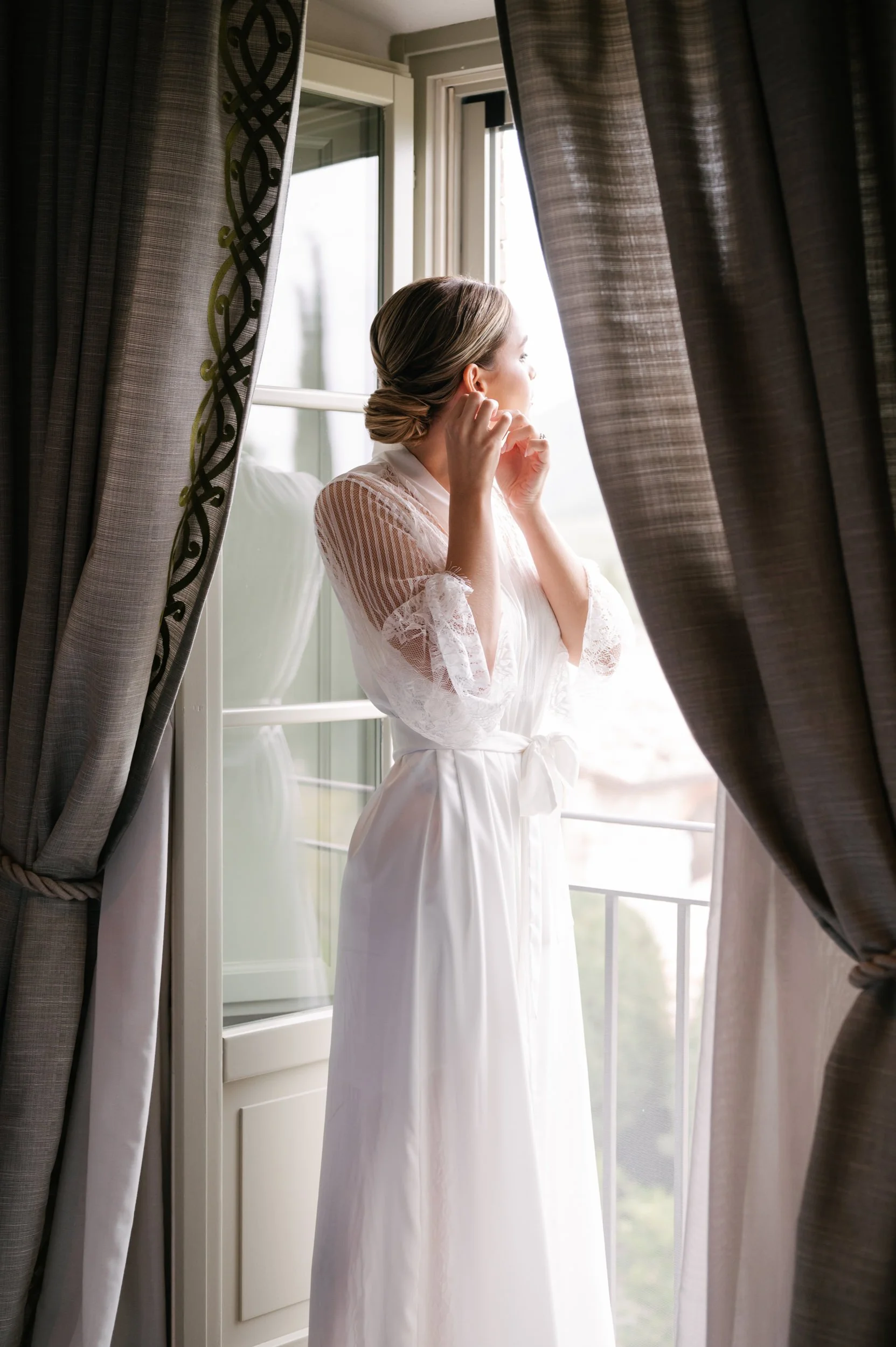 A woman in a white robe standing at an open window, adjusting her earrings, with dark curtains framing her.