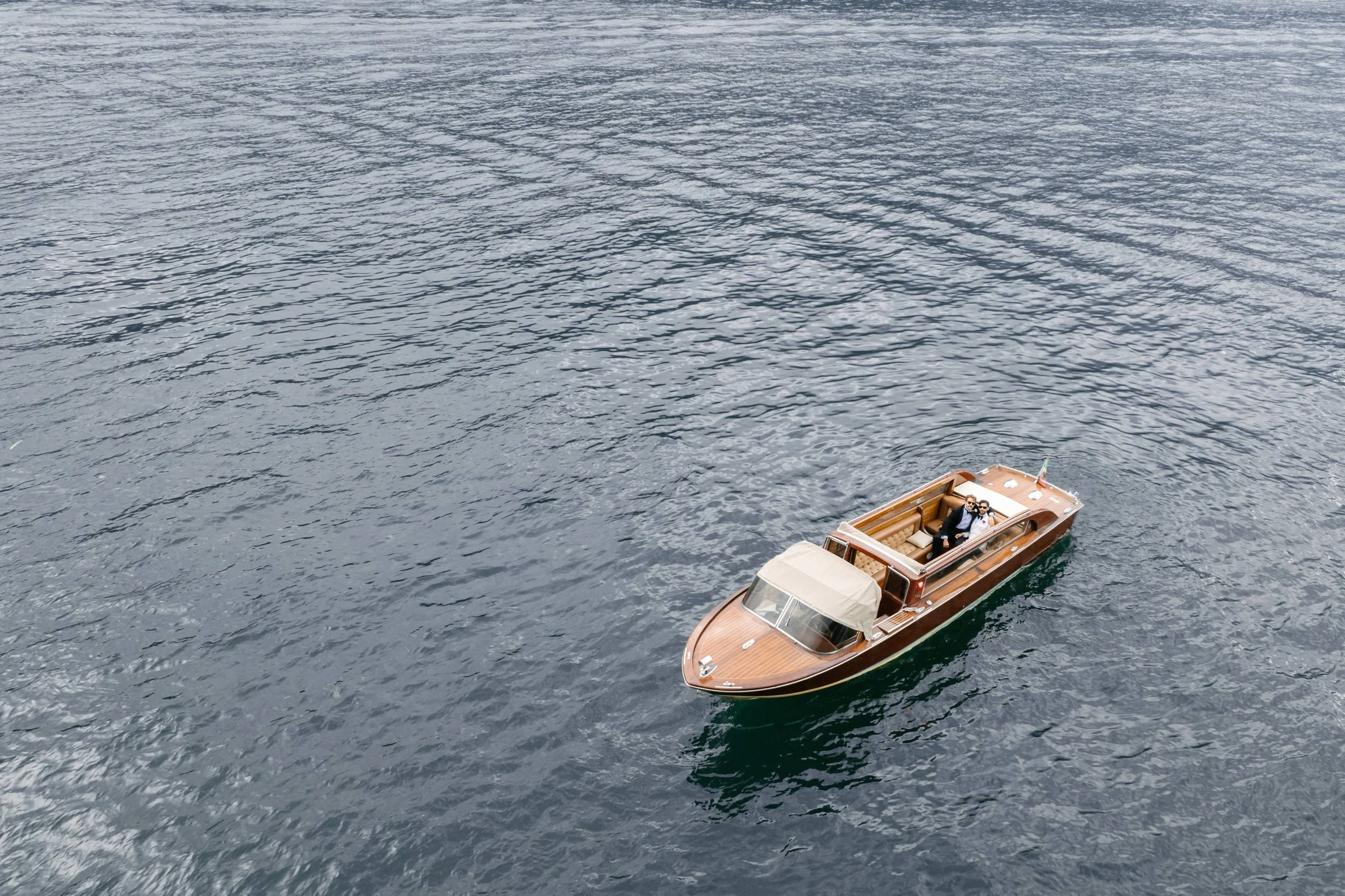 A small wooden boat with a cabin and an open deck, carrying two people, on calm water from an aerial perspective.
