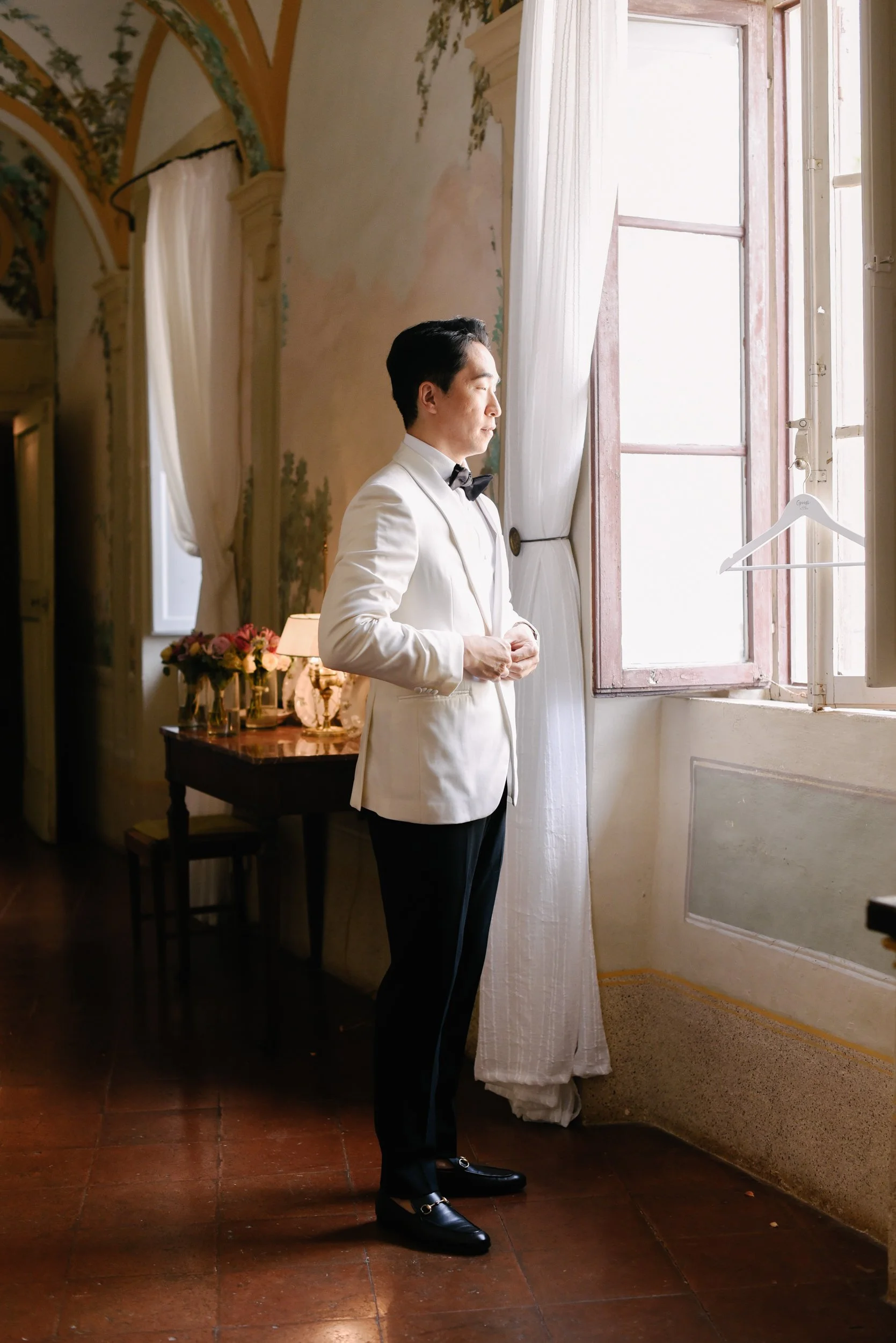A man dressed in a white tuxedo jacket with black pants and shoes, standing by an open window, looking outside with sunlight illuminating his face in an elegant room with floral decorations and vintage furniture.