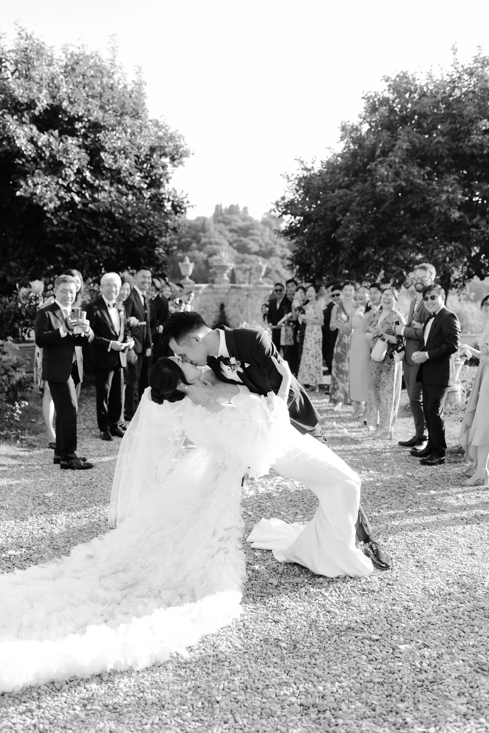 A black and white photo of a wedding celebration featuring a bride and groom in the foreground, with the groom dipping the bride as they share a kiss. Guests are gathered around, smiling and capturing the moment, outdoors with trees and decorative ur