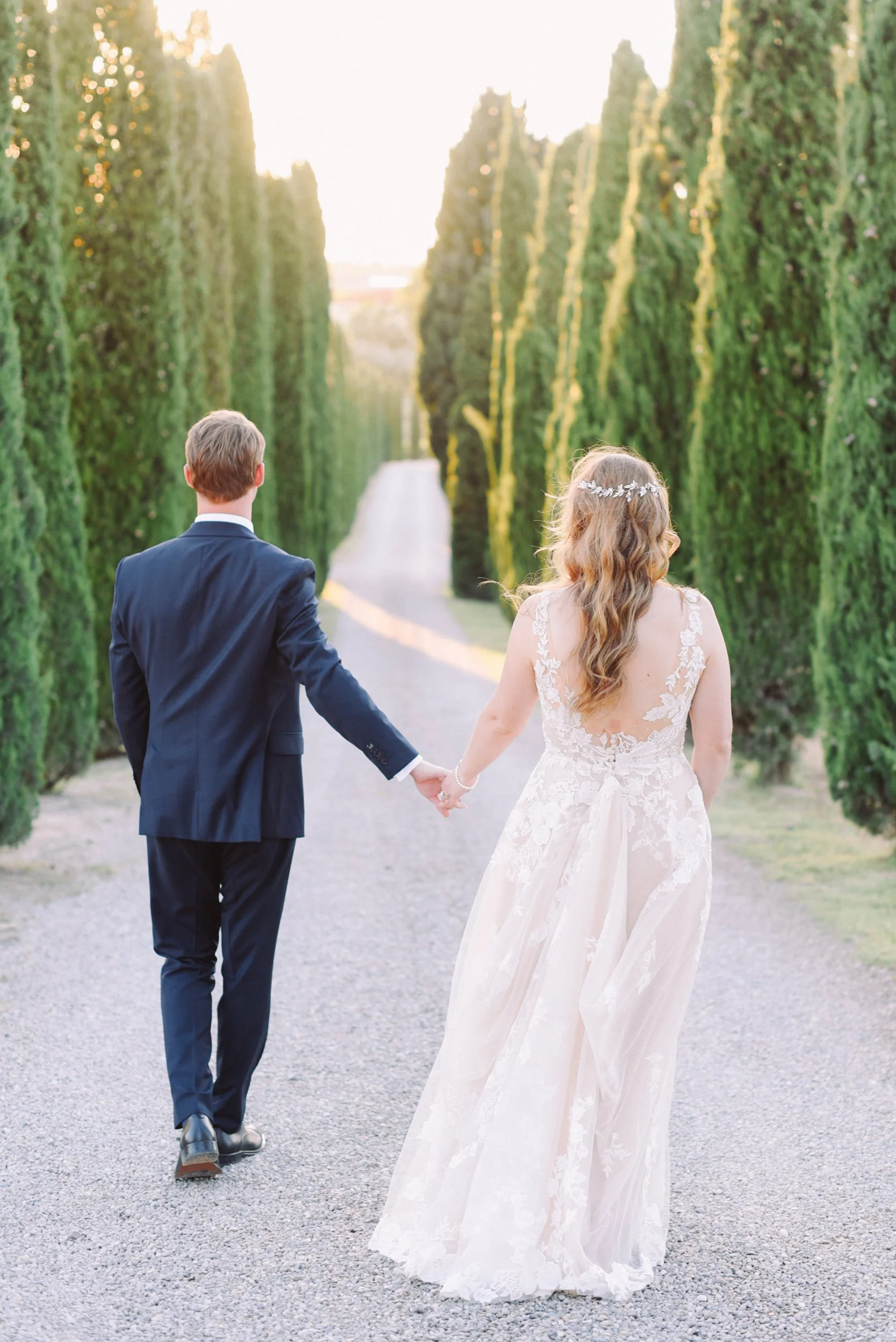 A bride and groom holding hands while walking down a gravel path lined with tall, green trees during sunset or sunrise.